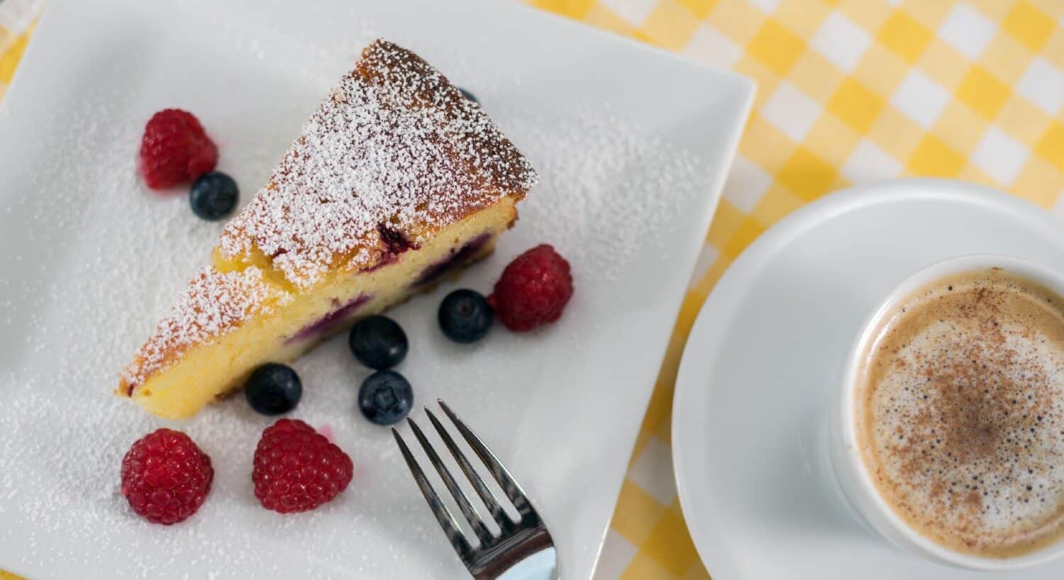 Slice of cake dusted with powdered sugar with berries on a white plate next to a cup of coffee