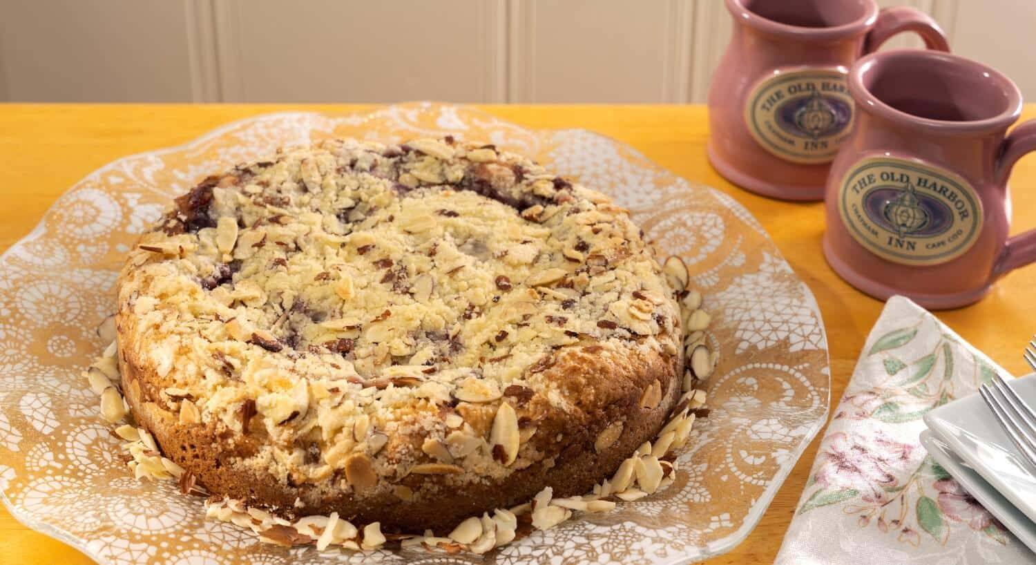 Golden brown round cake covered with slivered almonds next to two pink coffee mugs