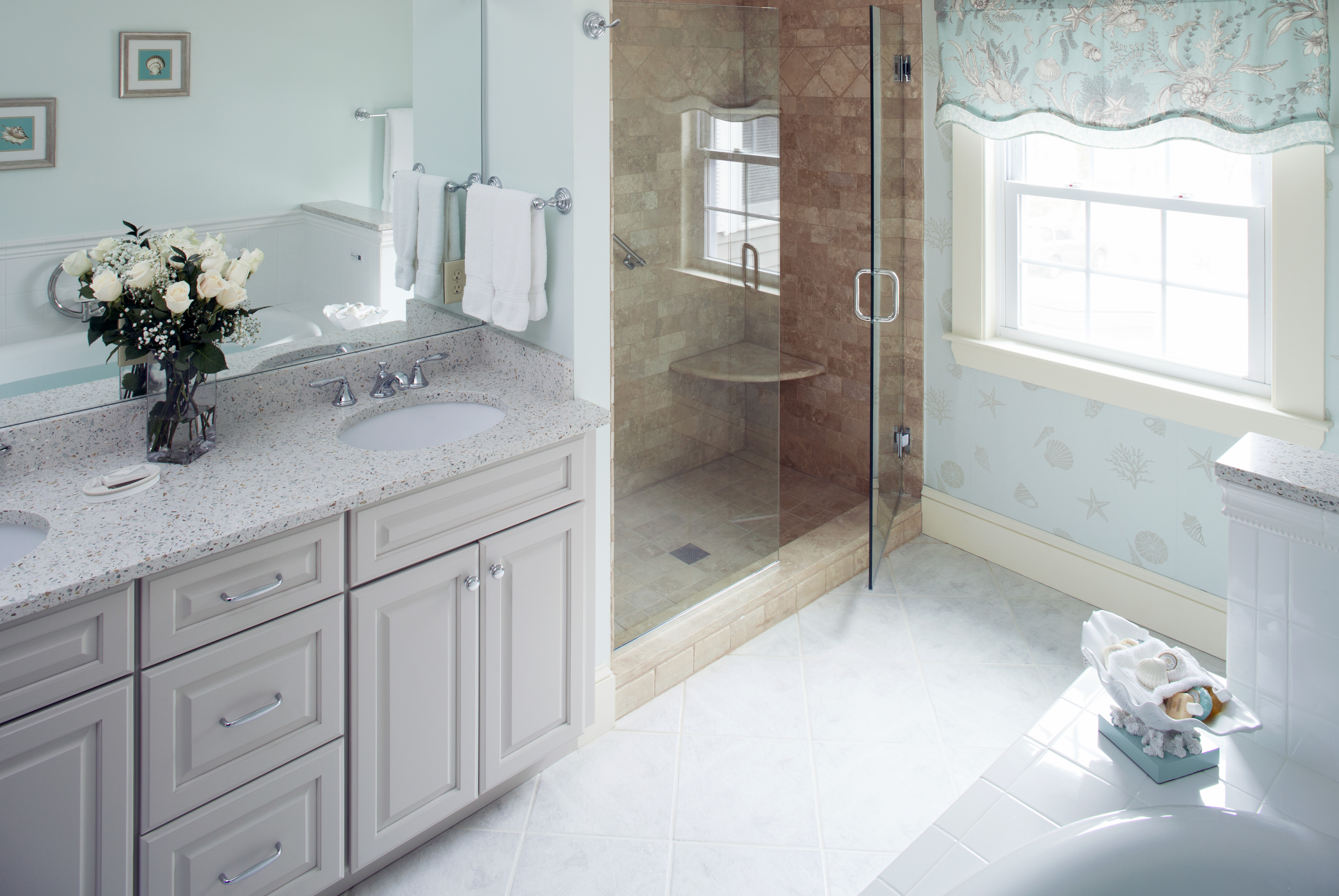 A large, elegant bathroom with a double vanity and a glass shower. The room has light-blue decor, a window letting in natural light, and a bouquet of white roses on the counter.