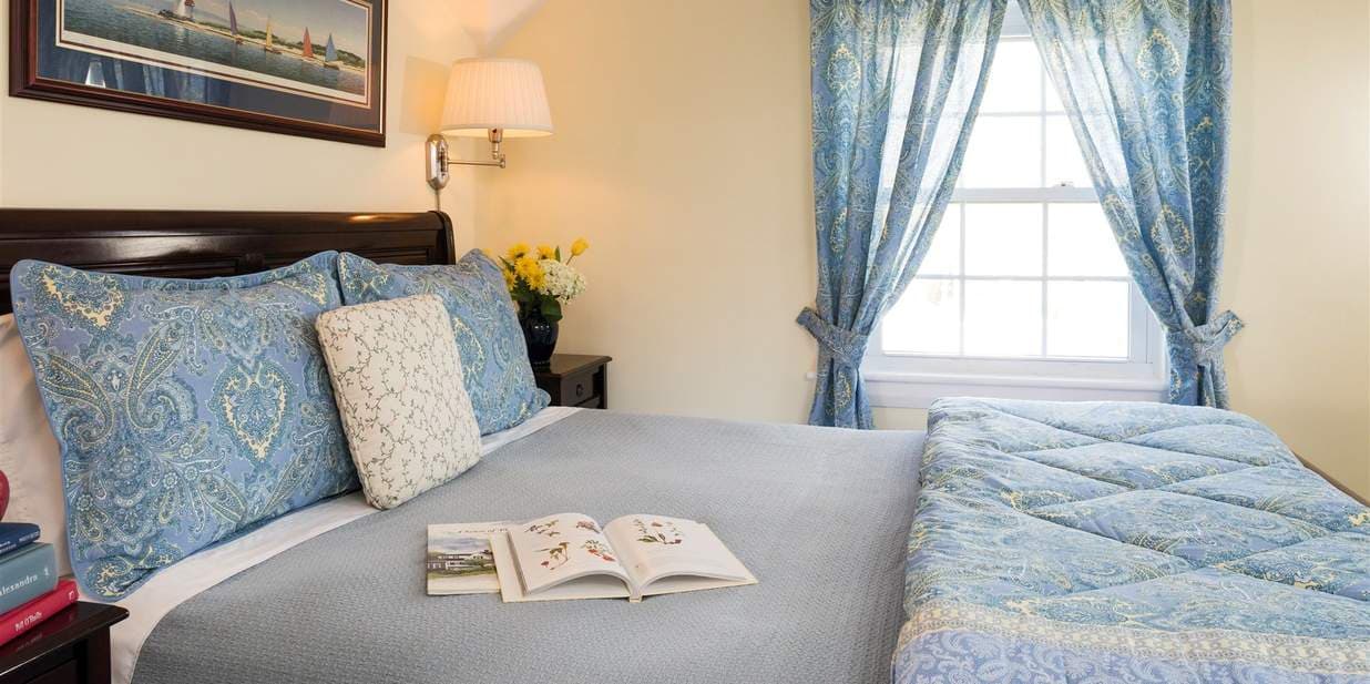 A comfortable bedroom with light yellow walls and a dark wood queen bed frame. The bed has blue pillows and a gray comforter, and an additional smaller bed with a blue comforter is next to it.