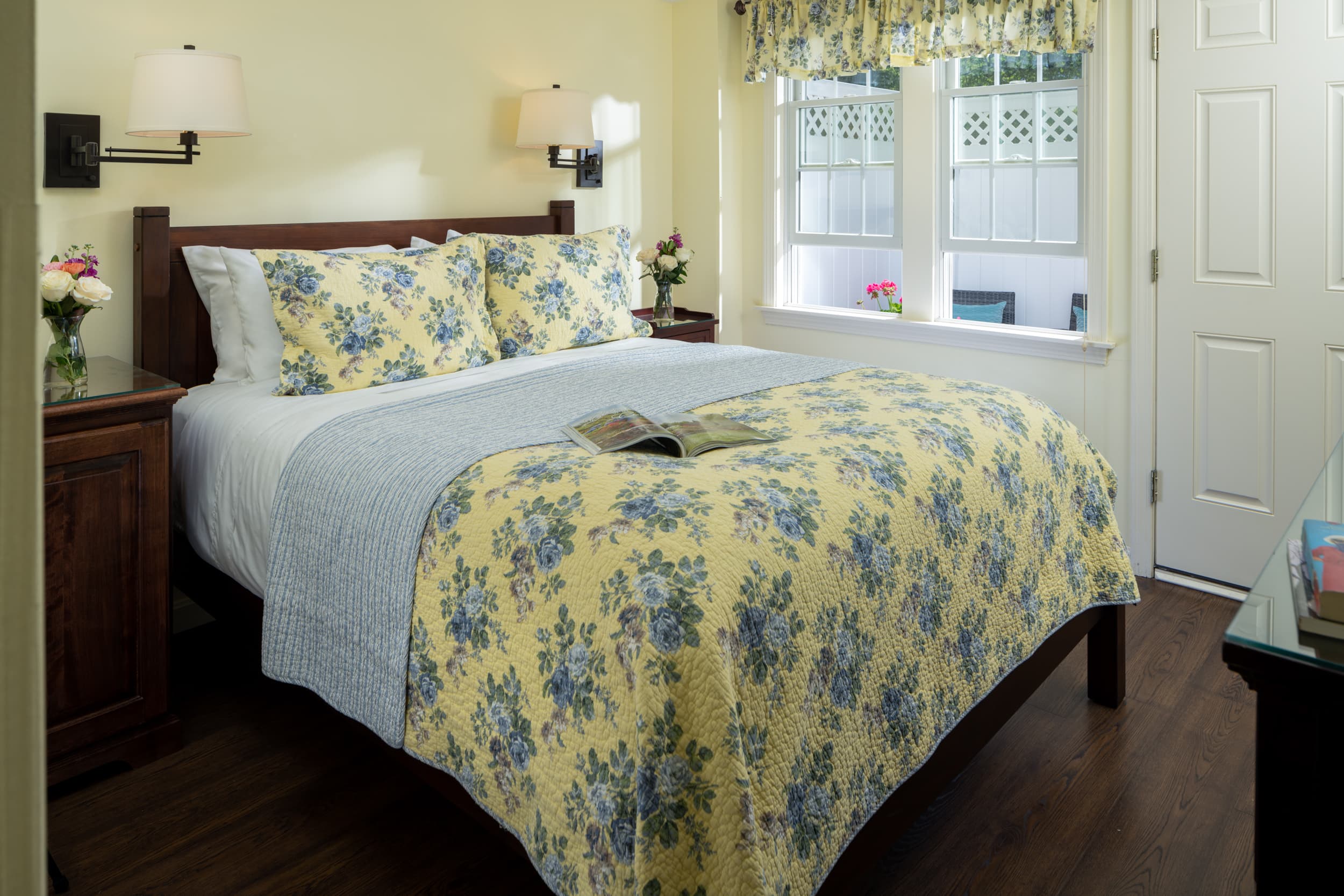 A charming bedroom with light yellow walls and a dark wood queen bed. The bed is dressed with a blue and yellow floral comforter, and the room has two bedside lamps, a window, and a private entrance.