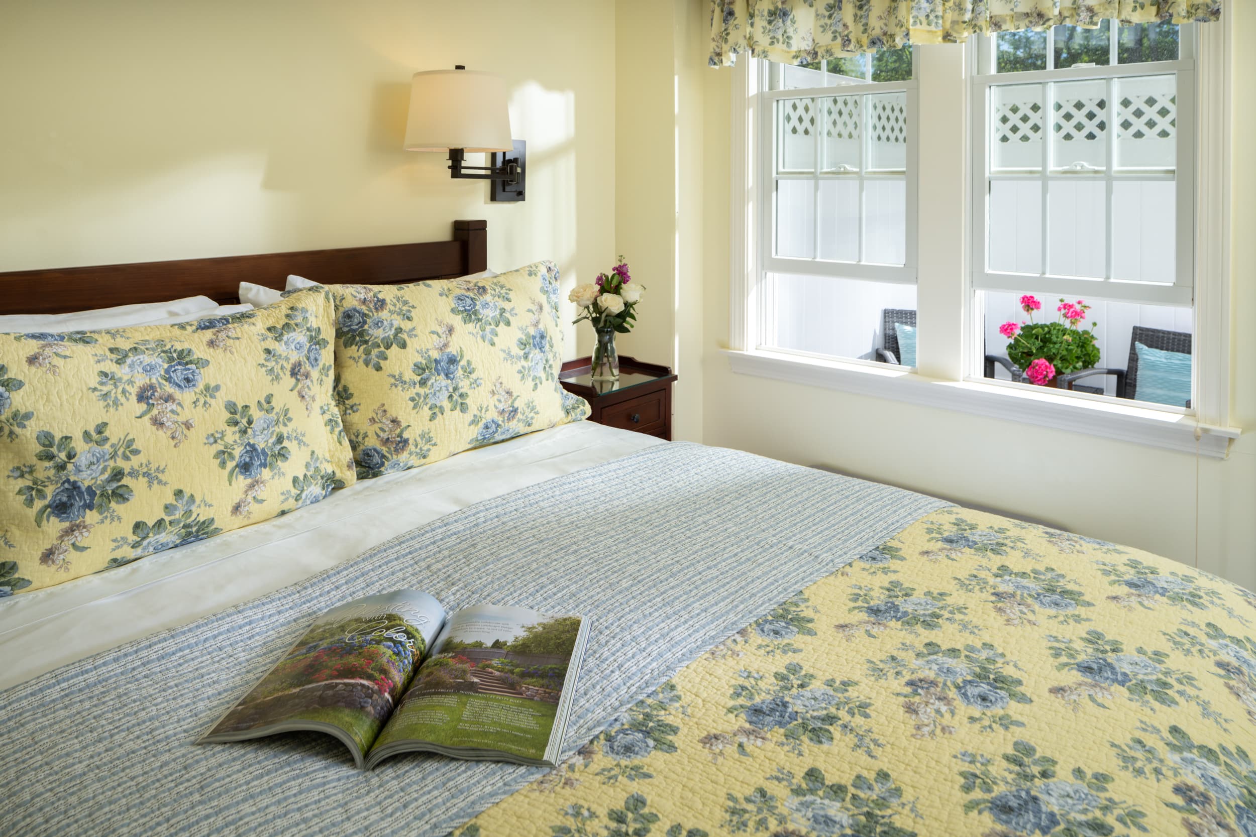 A peaceful bedroom with light yellow walls and a dark wood queen bed. The bed is dressed with a blue and yellow floral comforter, and the room has two bedside lamps, a window, and a private entrance.