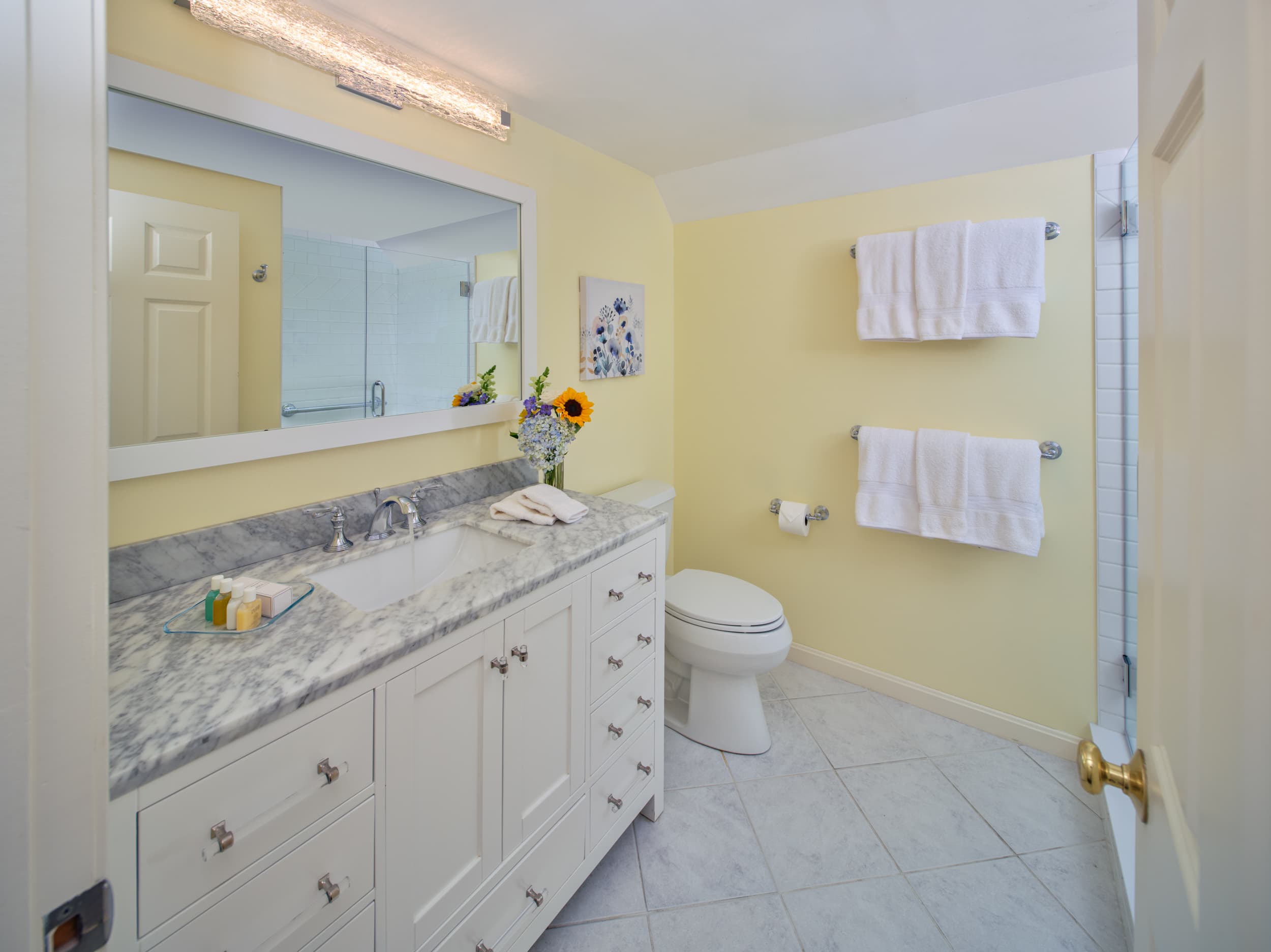 A bathroom with light yellow walls and a white vanity. The vanity has a white top and a single sink with a chrome faucet. A toilet is visible on the right, and a fresh towel is hanging on the door.