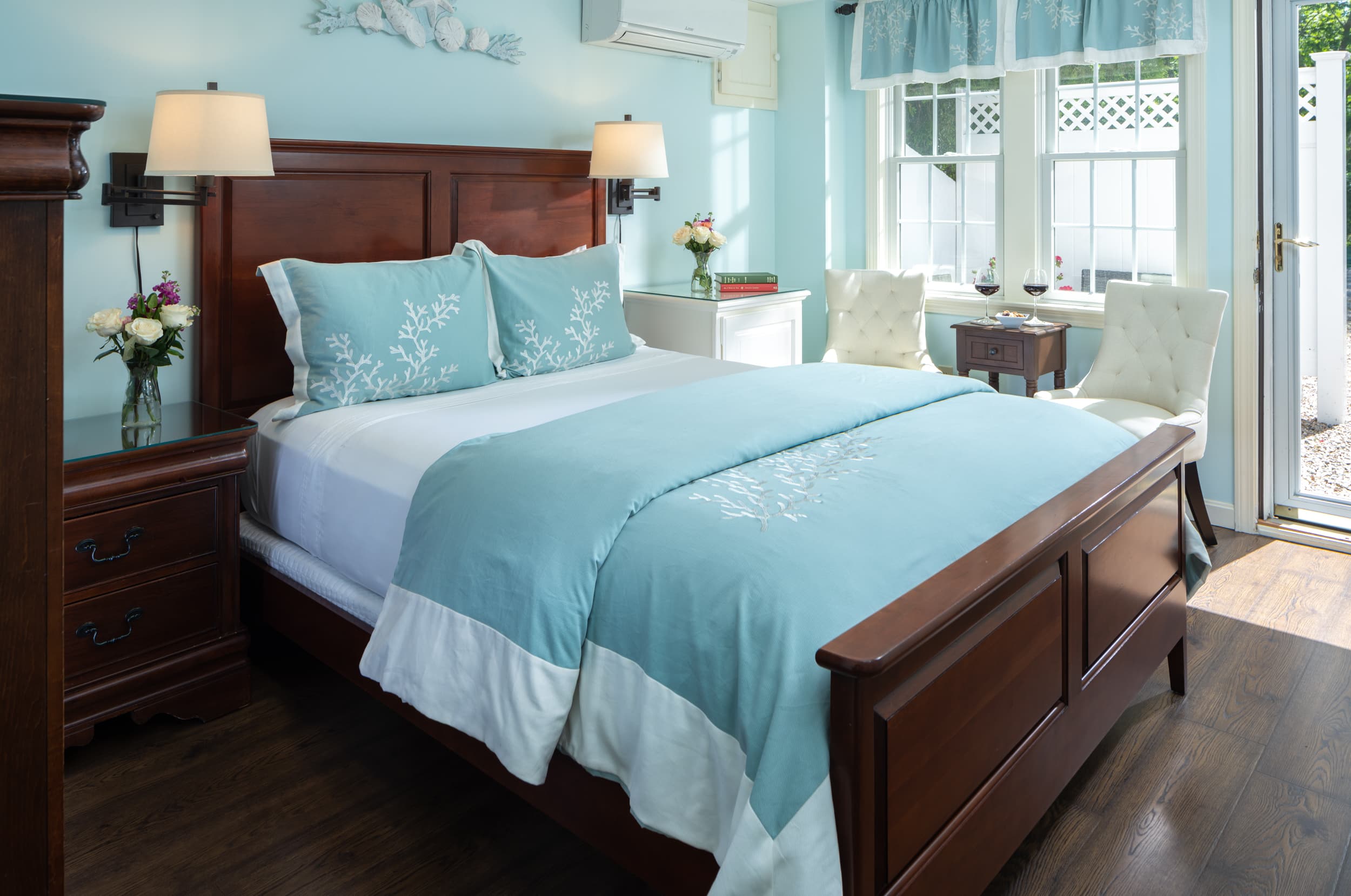 A light and airy bedroom with light blue walls and a dark wood bed frame. The bed is dressed with a blue comforter and pillows. Two tufted chairs sit in a sunny corner near a large window and a private entrance.