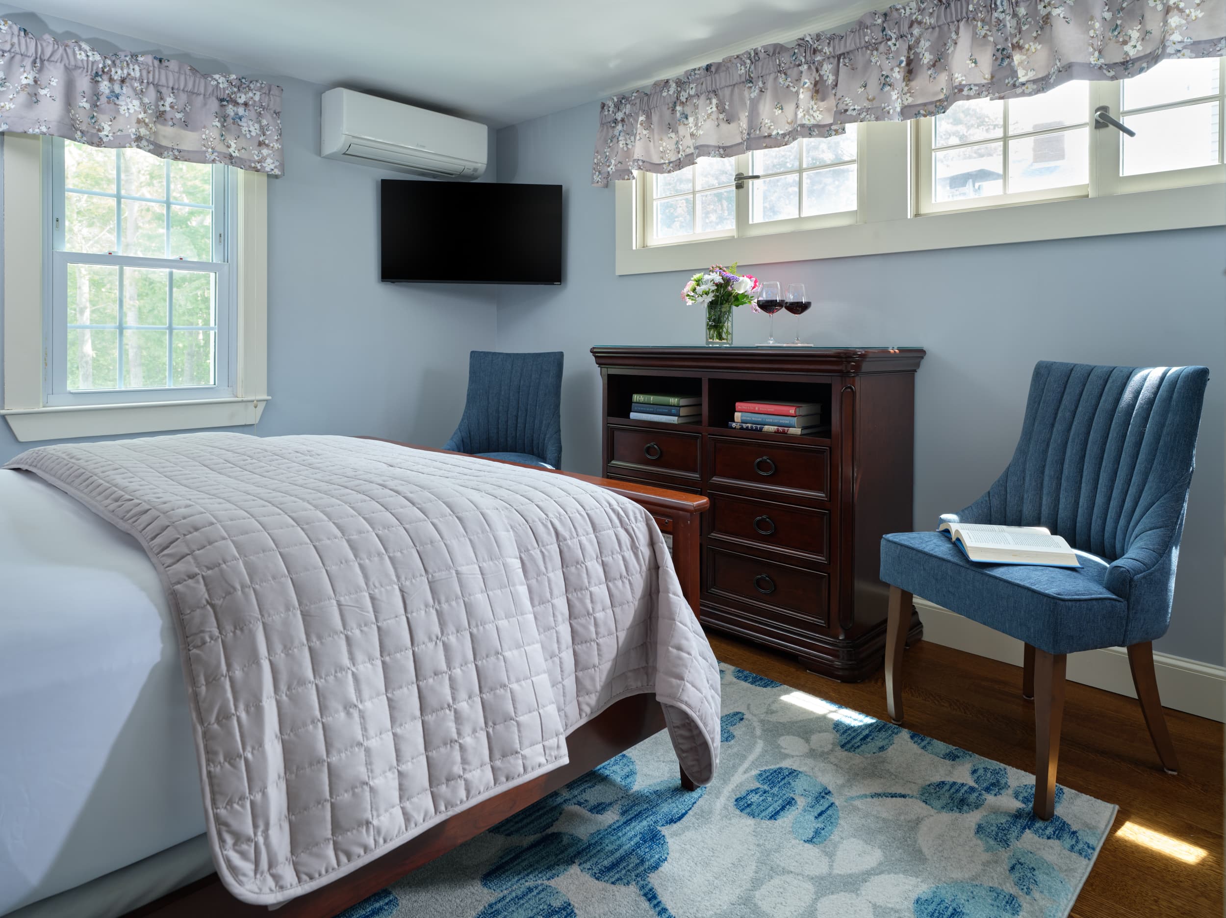 A peaceful bedroom with light blue walls, a dark wood dresser, and a flat-screen TV mounted on the wall. Two blue chairs with a book on one of them sit on a floral blue and white rug.