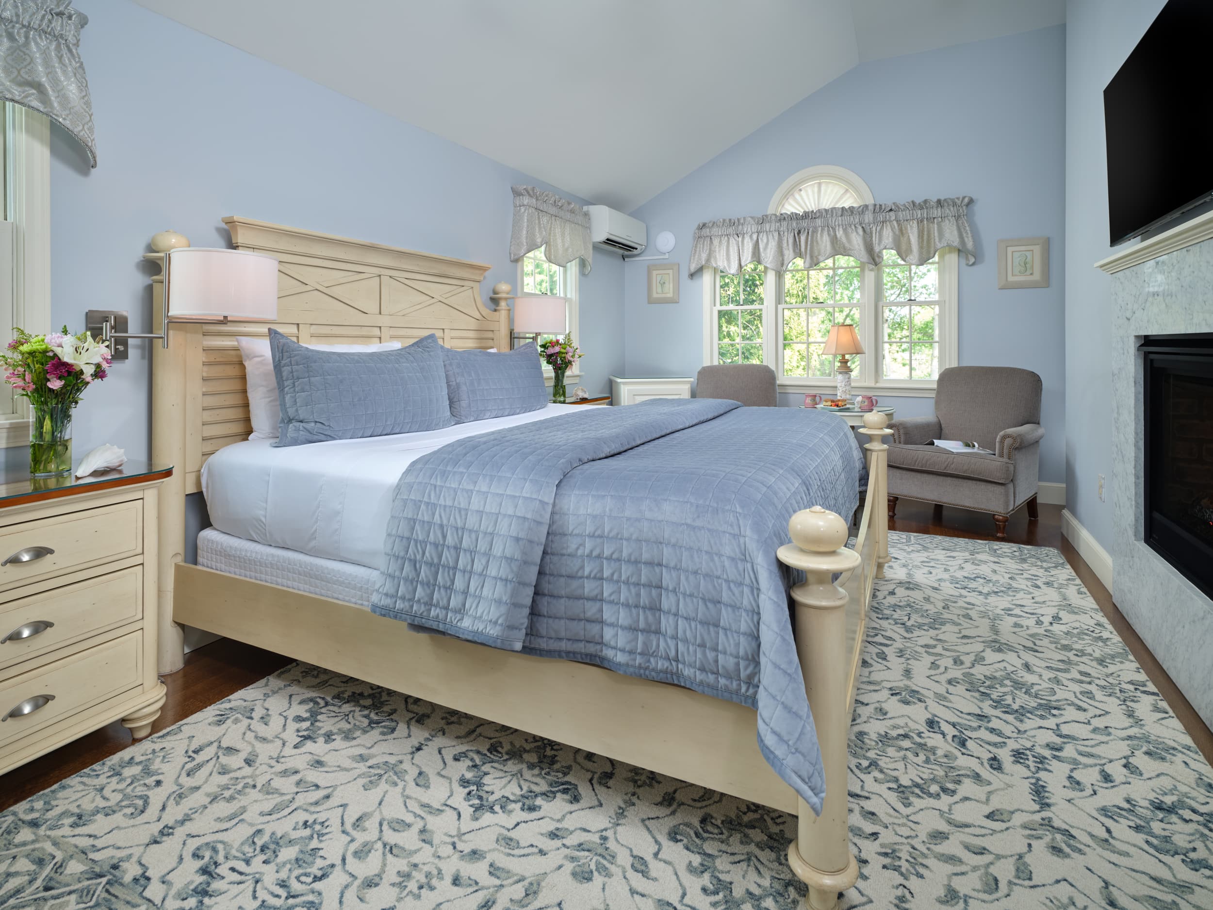 A tranquil bedroom with soft blue walls and cathedral ceilings. The room features a king-size bed, a fireplace, and a large window with natural light.