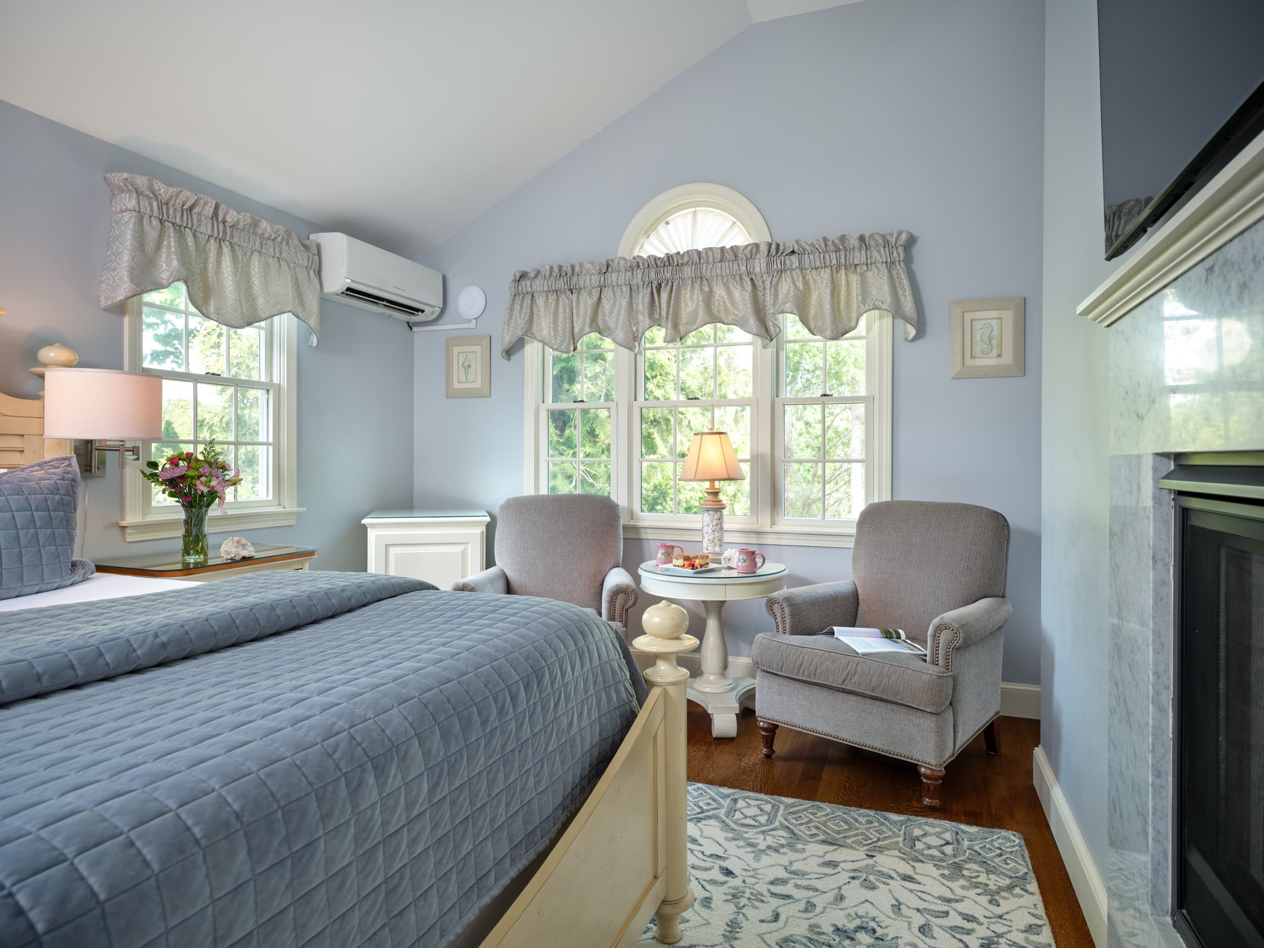 A peaceful bedroom with light blue walls and a vaulted ceiling. Two armchairs and a small table sit by a window with a view of lush greenery.
