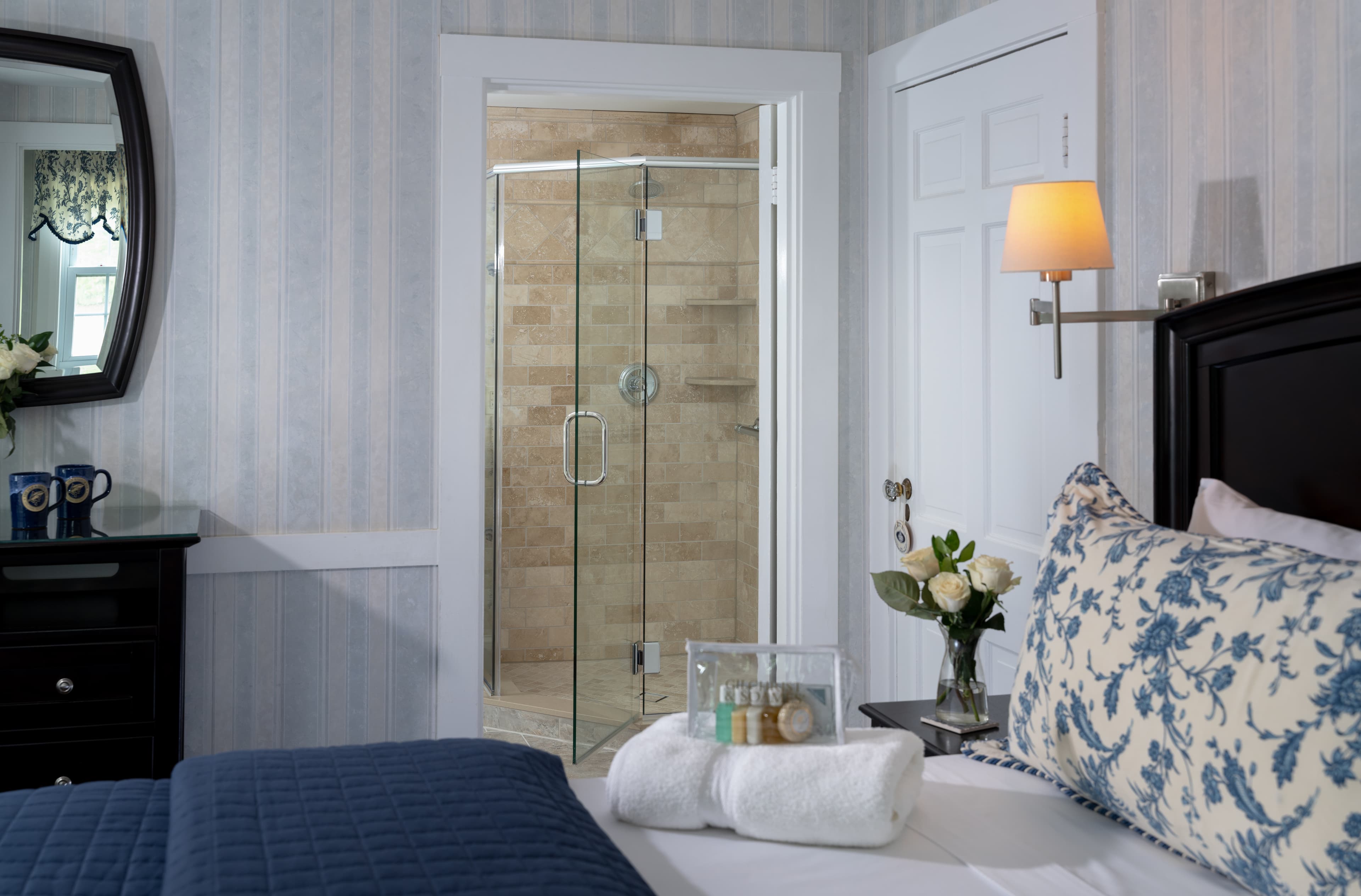 A cozy bedroom with blue and white decor. The bed is made with a blue comforter and a decorative pillow. The ensuite bathroom with a glass shower is visible in the background.