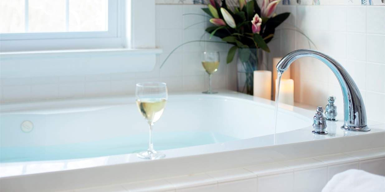 A tranquil bathroom scene with a wine glass, bouquet of flowers, & candles on the edge of a Jacuzzi tub.