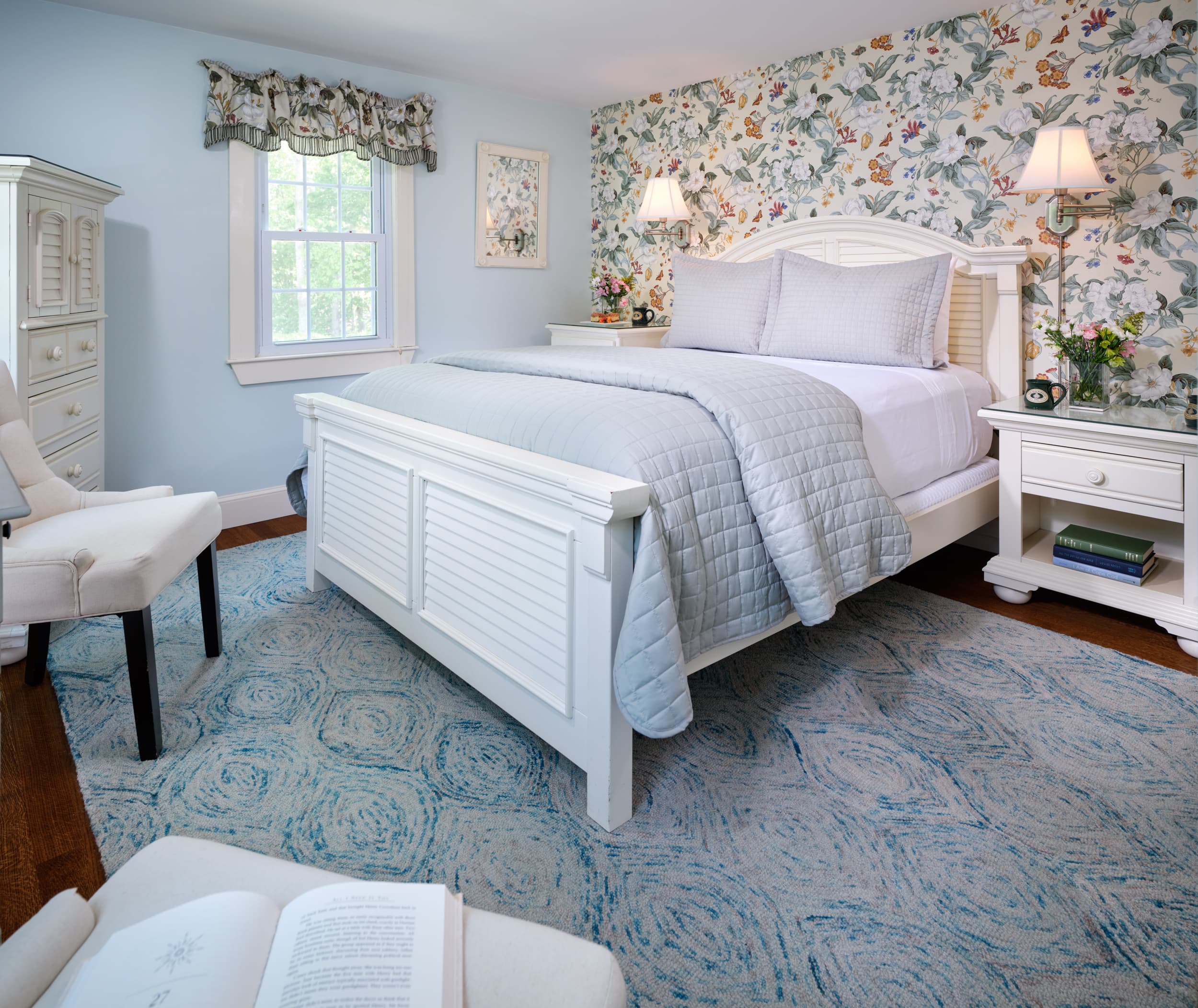 A light and airy bedroom with light-blue walls and a white floral accent wall. The room has a white queen bed with blue bedding, a blue and white patterned rug, and a white chaise lounge.