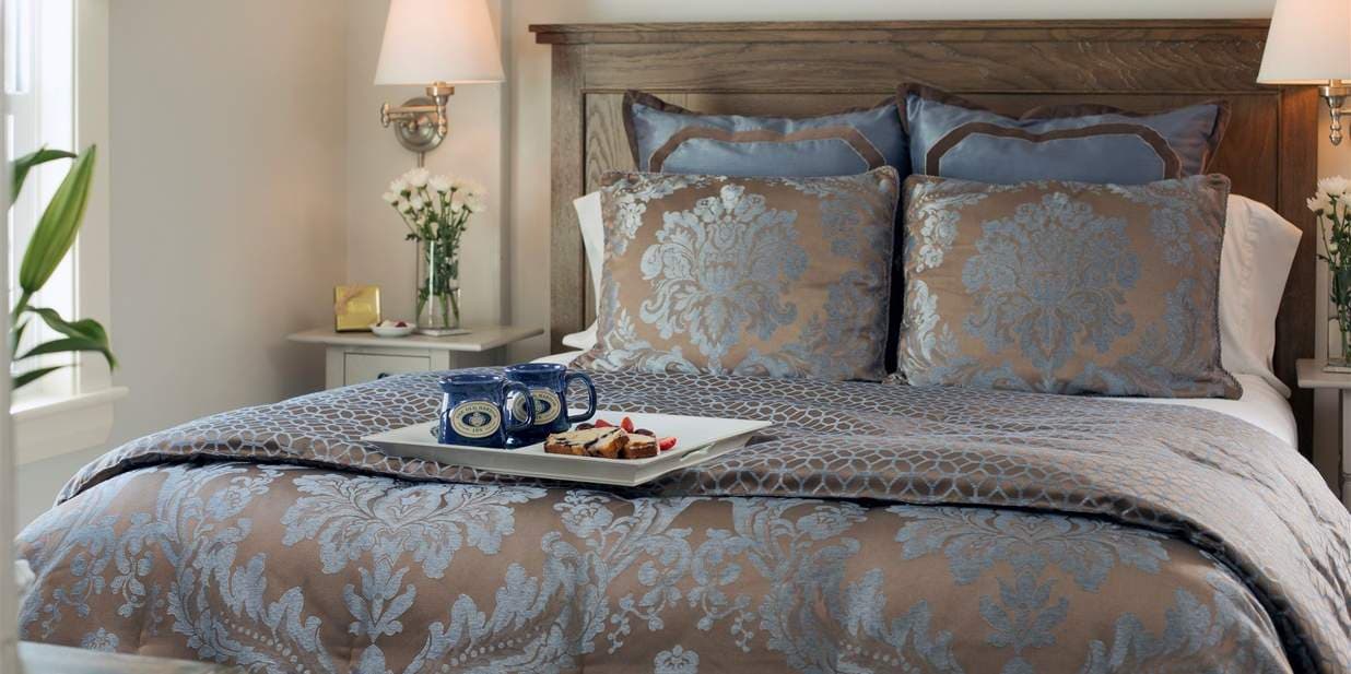 A queen bed with a gray and blue floral comforter and pillows. A tray with two mugs and snacks is on the comforter, and lamps on nightstands illuminate the room.