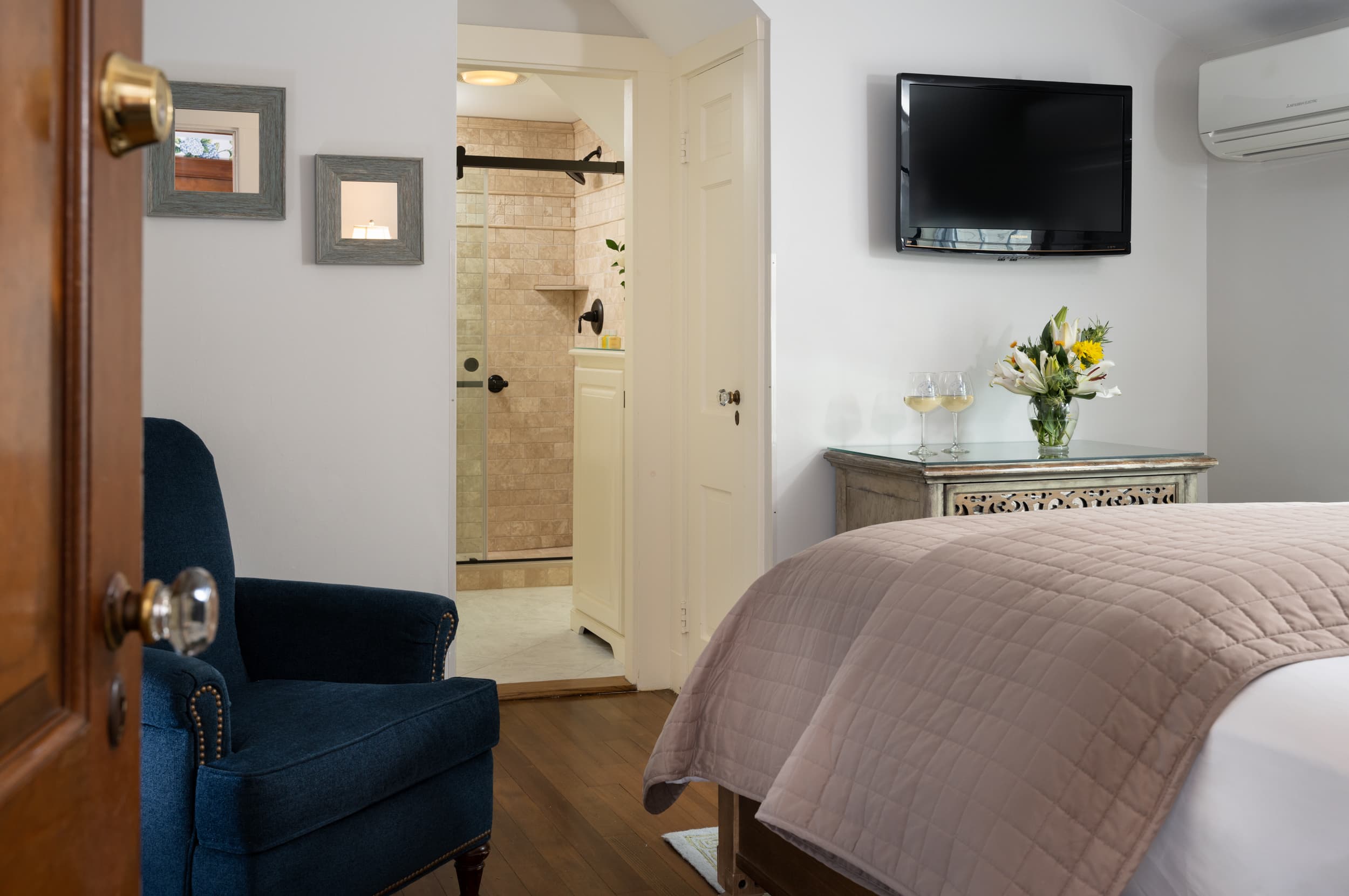 A comfortable bedroom with an open door to a bathroom. The bedroom has a dark blue armchair and a queen bed with a light pink comforter. A flat-screen TV is mounted above a chest of drawers.