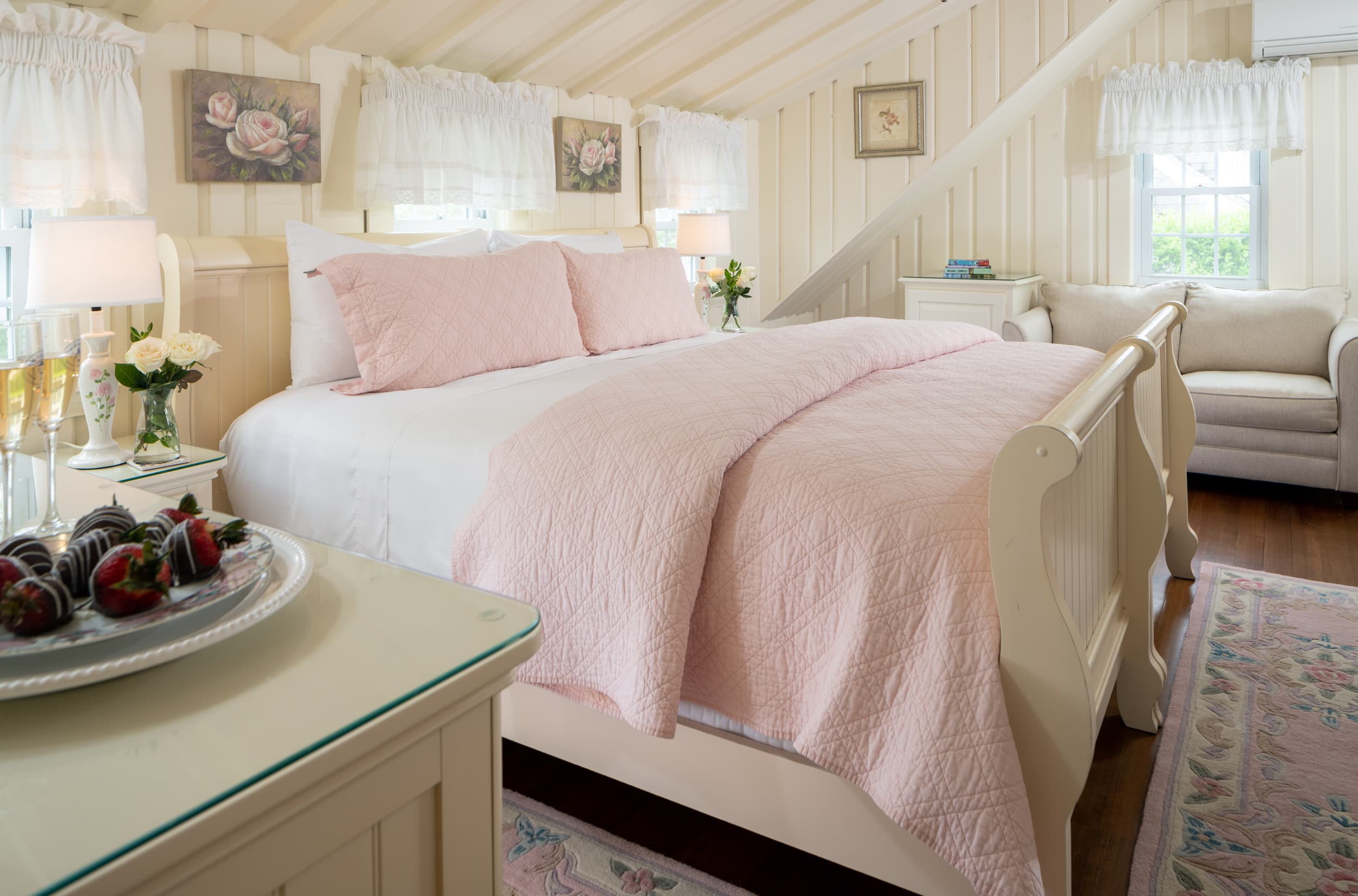 A soft and inviting bedroom with a sloped ceiling and a white sleigh bed. The room is decorated with shades of pink and white, with a tray of chocolate-covered strawberries.