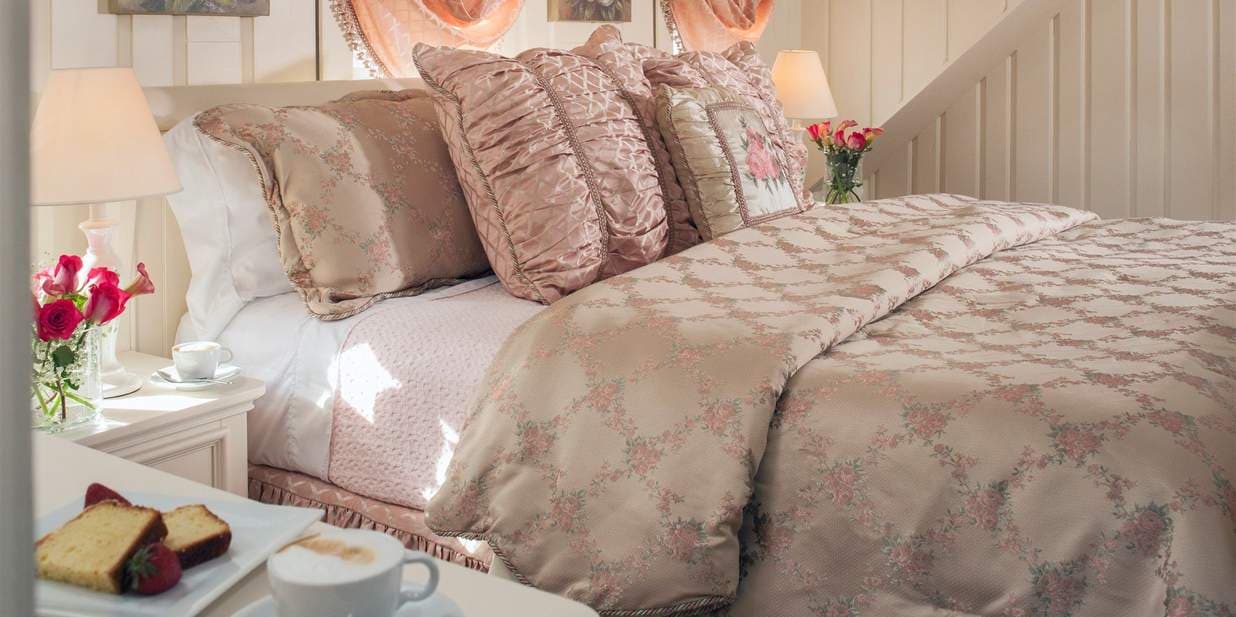 A serene bedroom with a bed covered in a muted pink quilt and pillows. A small vase of pink flowers and a tray with chocolates sit on a bedside table.