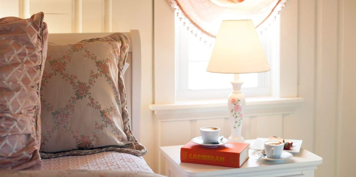 A soft, cozy bedroom scene with a pillow and a lamp on a bedside table. The lamp illuminates a book and two teacups, creating a relaxing atmosphere.