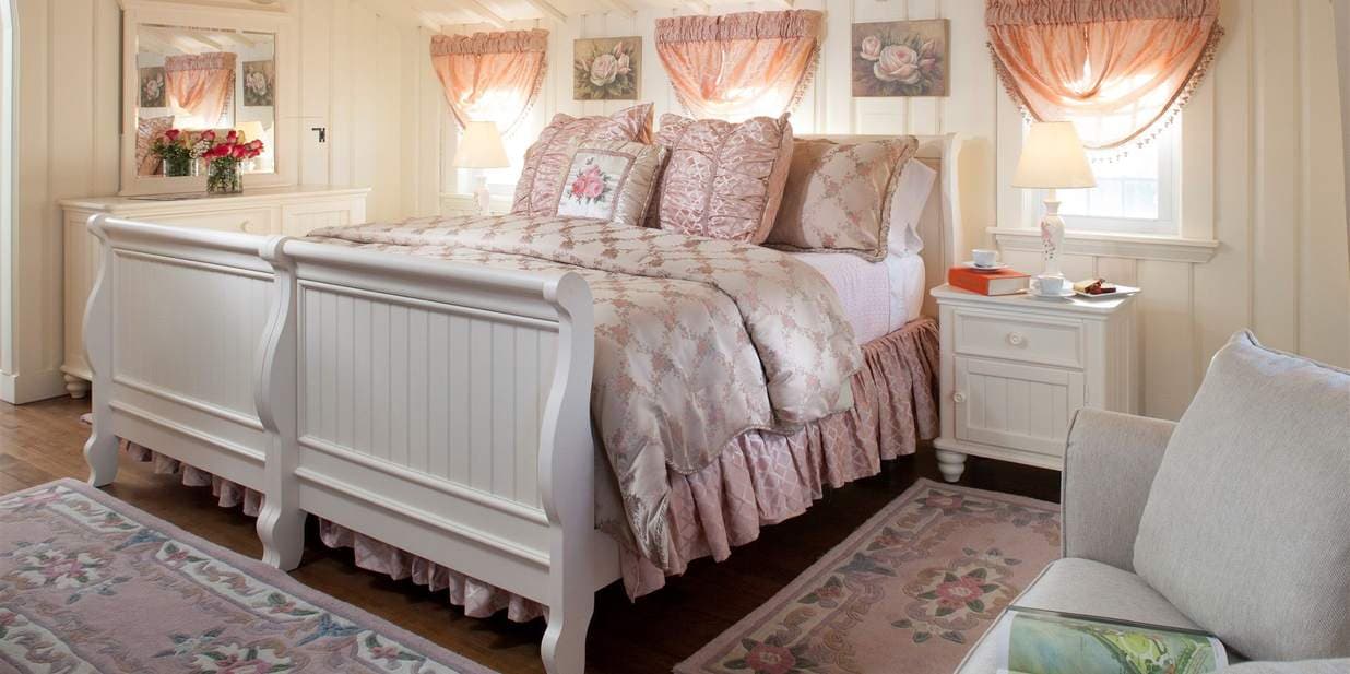 A light and elegant bedroom with two white twin beds pushed together. The beds are covered in pink floral bedding, and a soft pink rug is on the floor.