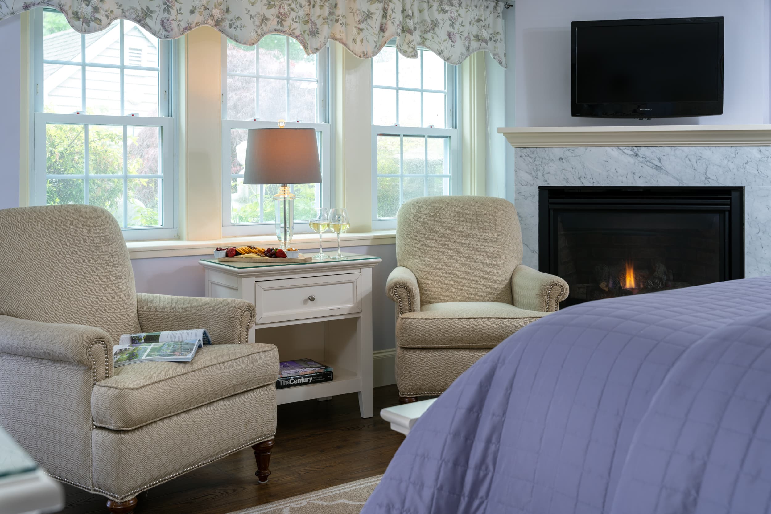 A serene sitting area within a bedroom, featuring two comfortable armchairs, a side table, and a lit fireplace with a TV mounted above it.