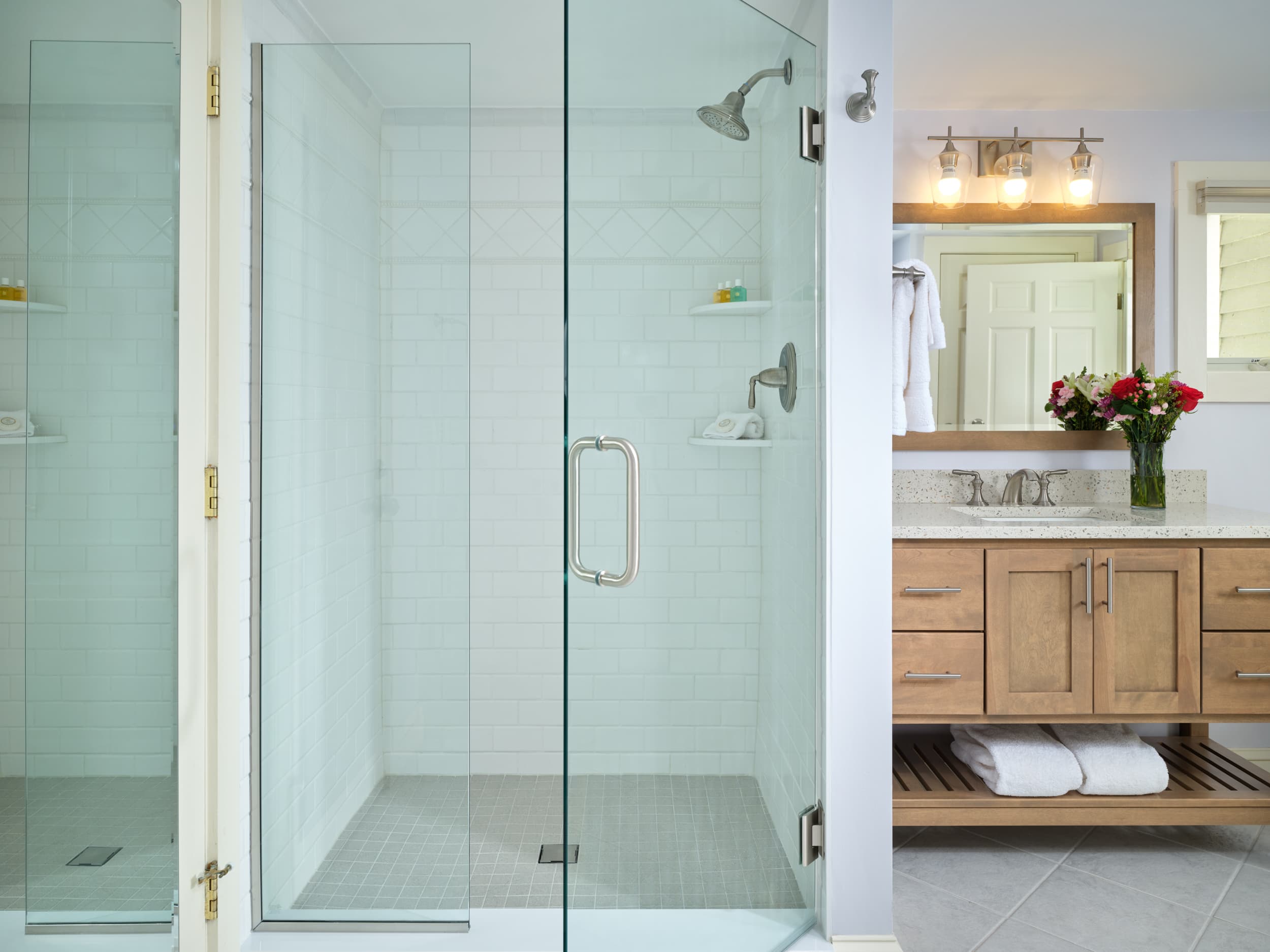 A large, bright luxury bathroom with a frameless glass shower and a double vanity. The vanity has a light wood finish and a white countertop with a vessel sink.