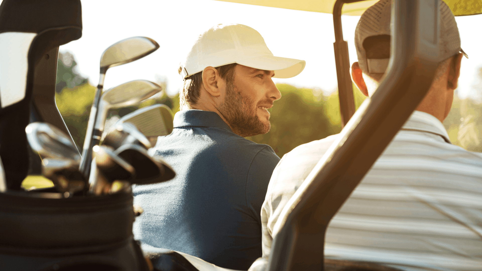 Two men in a golf cart, with golf clubs in the foreground, enjoying a sunny day on the course.