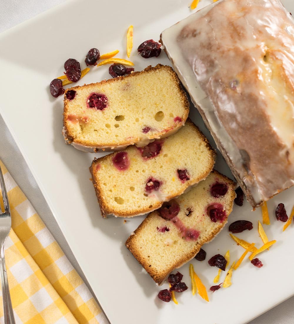 Slices of glazed cranberry-orange bread on a white plate, garnished with dried cranberries and citrus zest.