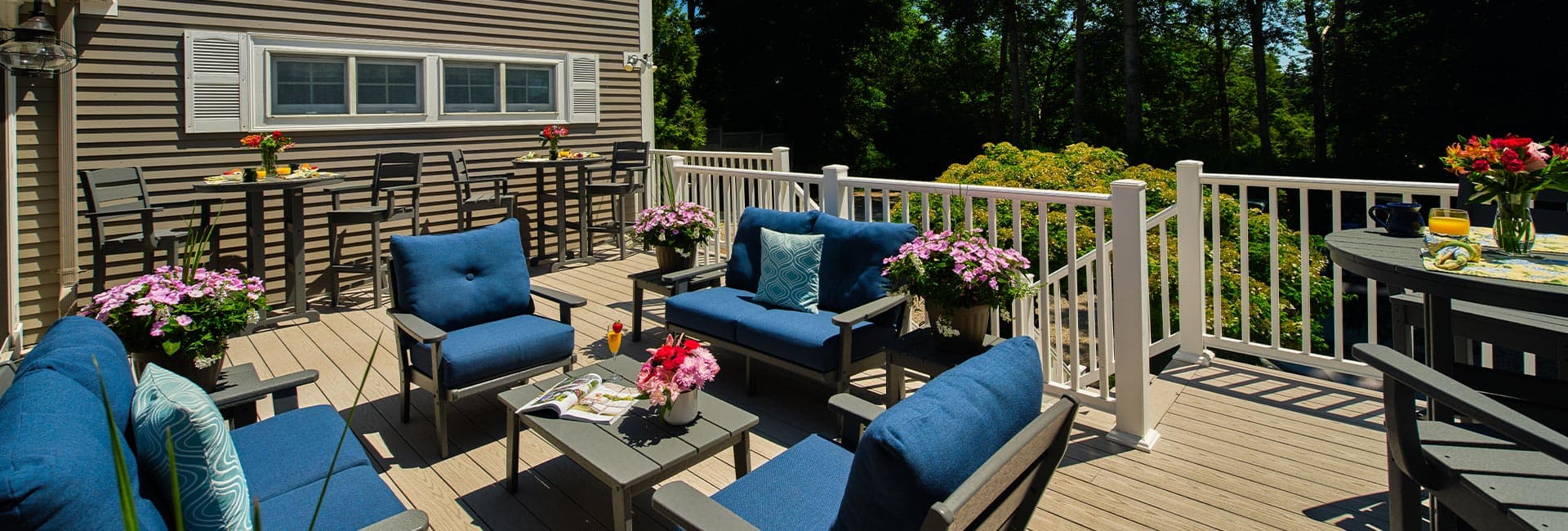 A cozy outdoor seating area with blue chairs, a coffee table, and flower pots surrounded by greenery.