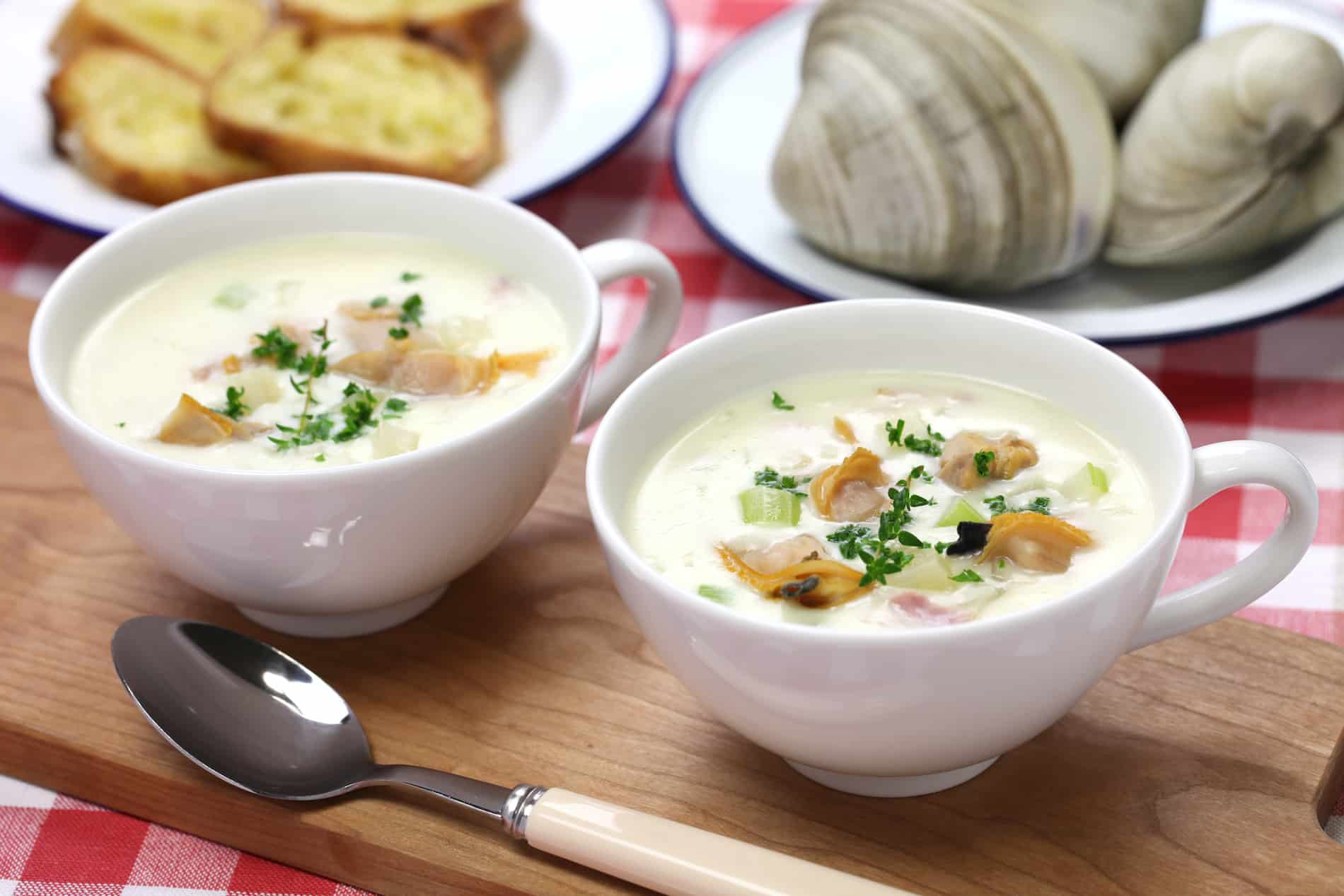 Two bowls of creamy clam chowder topped with herbs, accompanied by slices of garlic bread and clams.