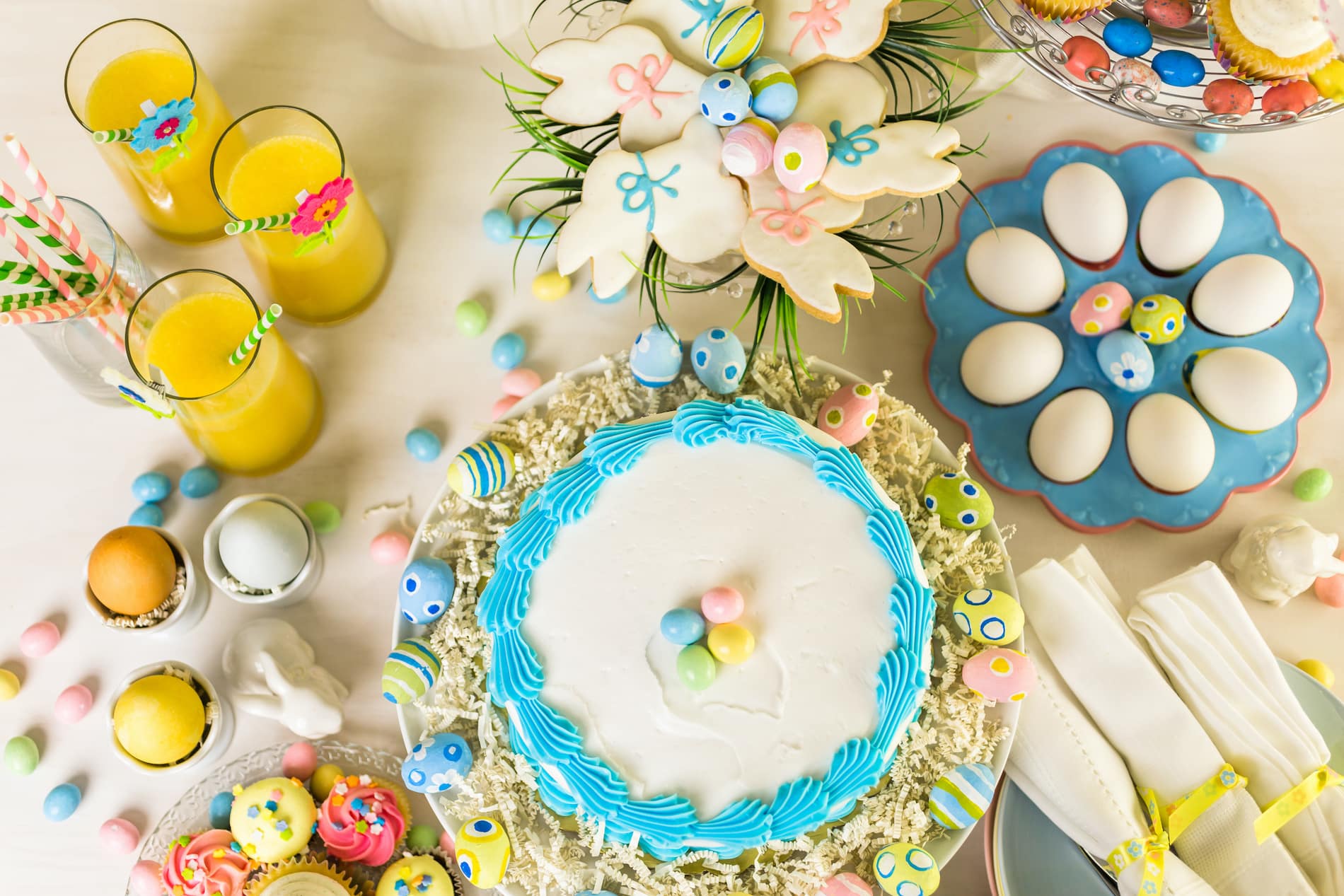 A colorful Easter dessert spread featuring a cake with blue icing, decorated cookies, cupcakes, colored eggs, and yellow drinks.