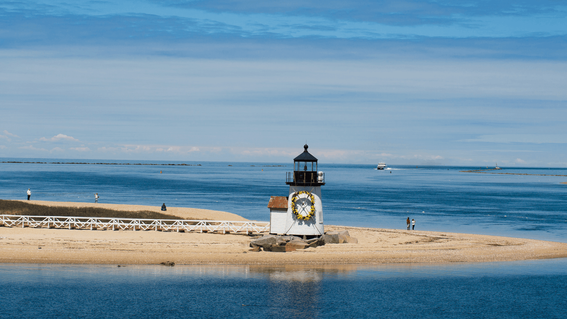 A lighthouse sits on a sandy shore, surrounded by calm waters and a blue sky.
