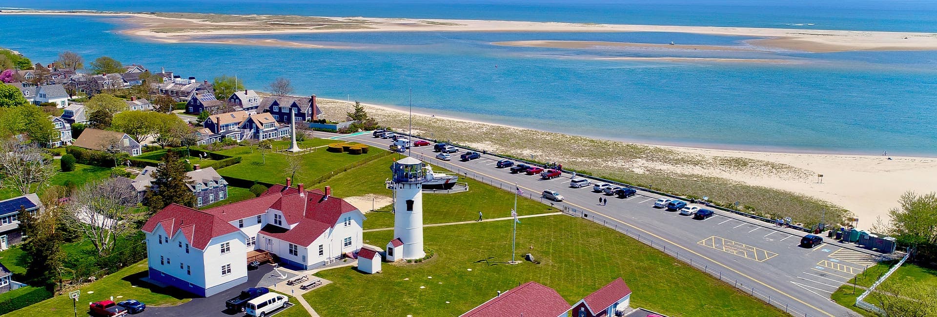 Aerial view of a coastal area featuring a lighthouse, green lawns, and residential homes by the beach.