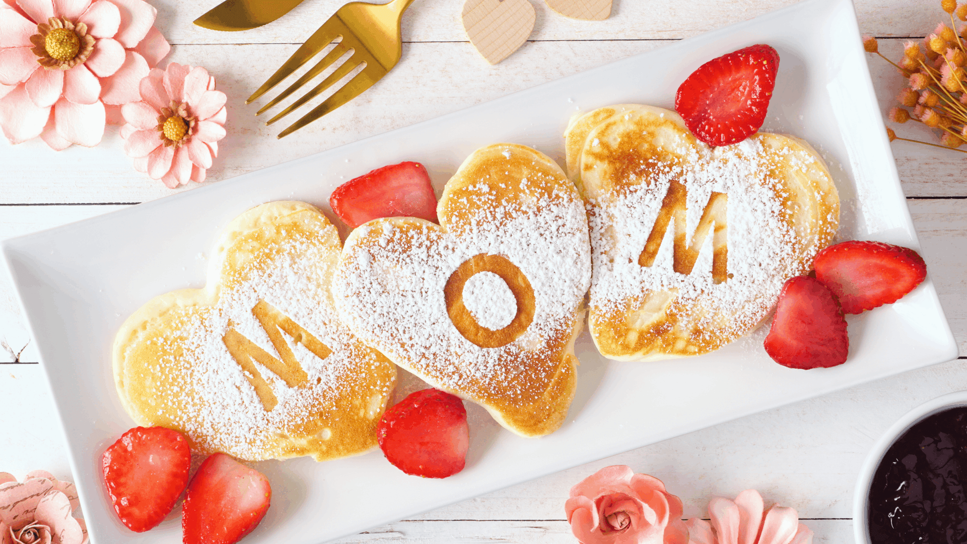 A plate of heart-shaped pancakes spells "MOM," surrounded by fresh strawberries and powdered sugar. A plate of heart-shaped pancakes spells "MOM," surrounded by fresh strawberries and powdered sugar.