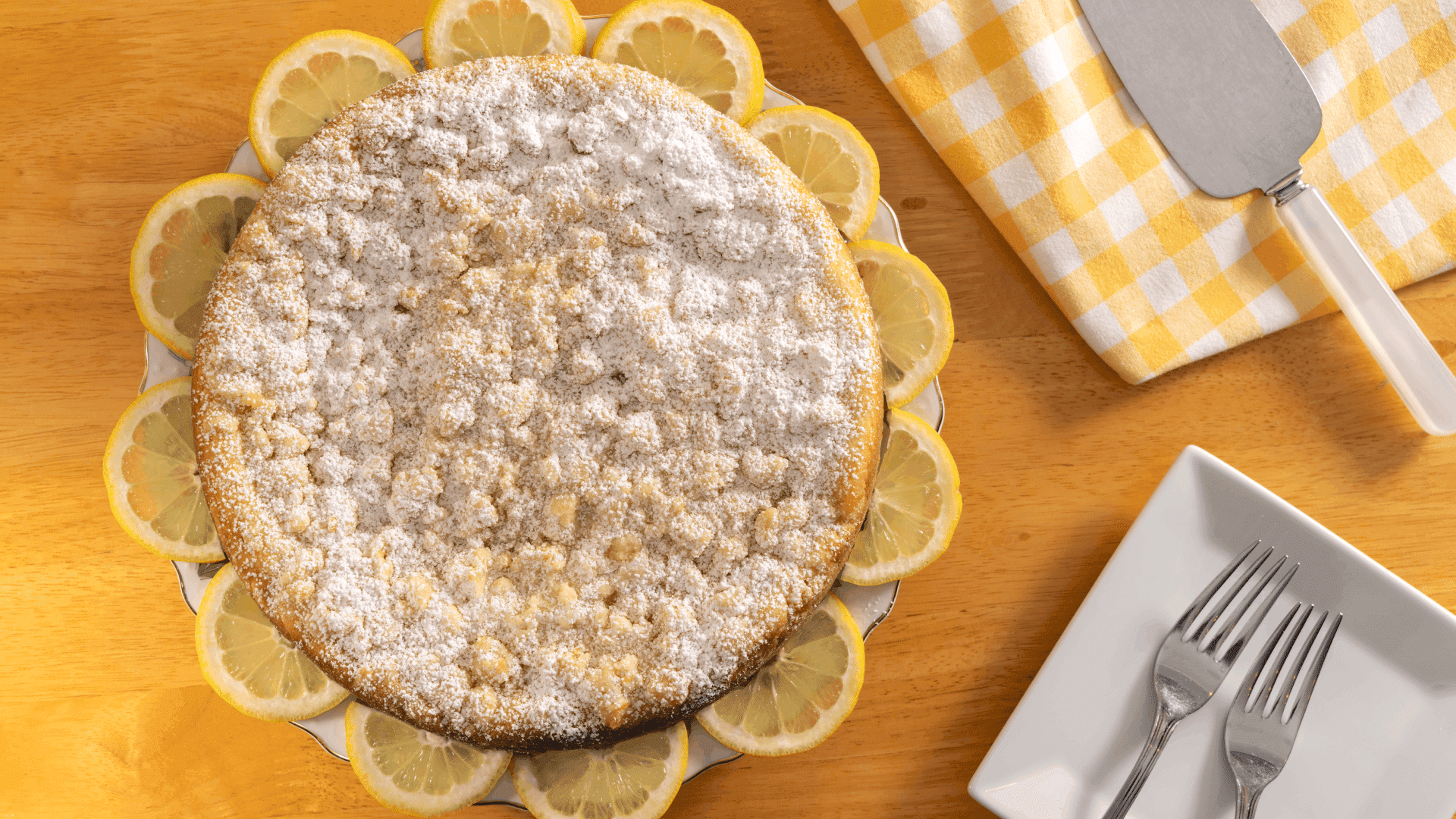 A lemon cake dusted with powdered sugar, surrounded by lemon slices, on a wooden table with utensils nearby.
