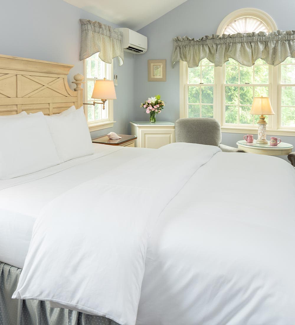 Bright and airy bedroom with a white bedspread, light blue walls, and a floral arrangement by the window.
