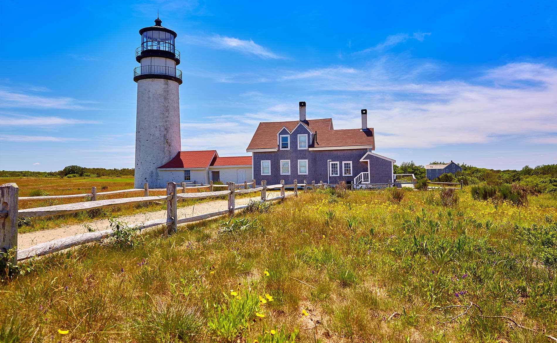 Lighthouse beside a scenic house in a grassy landscape under a blue sky.