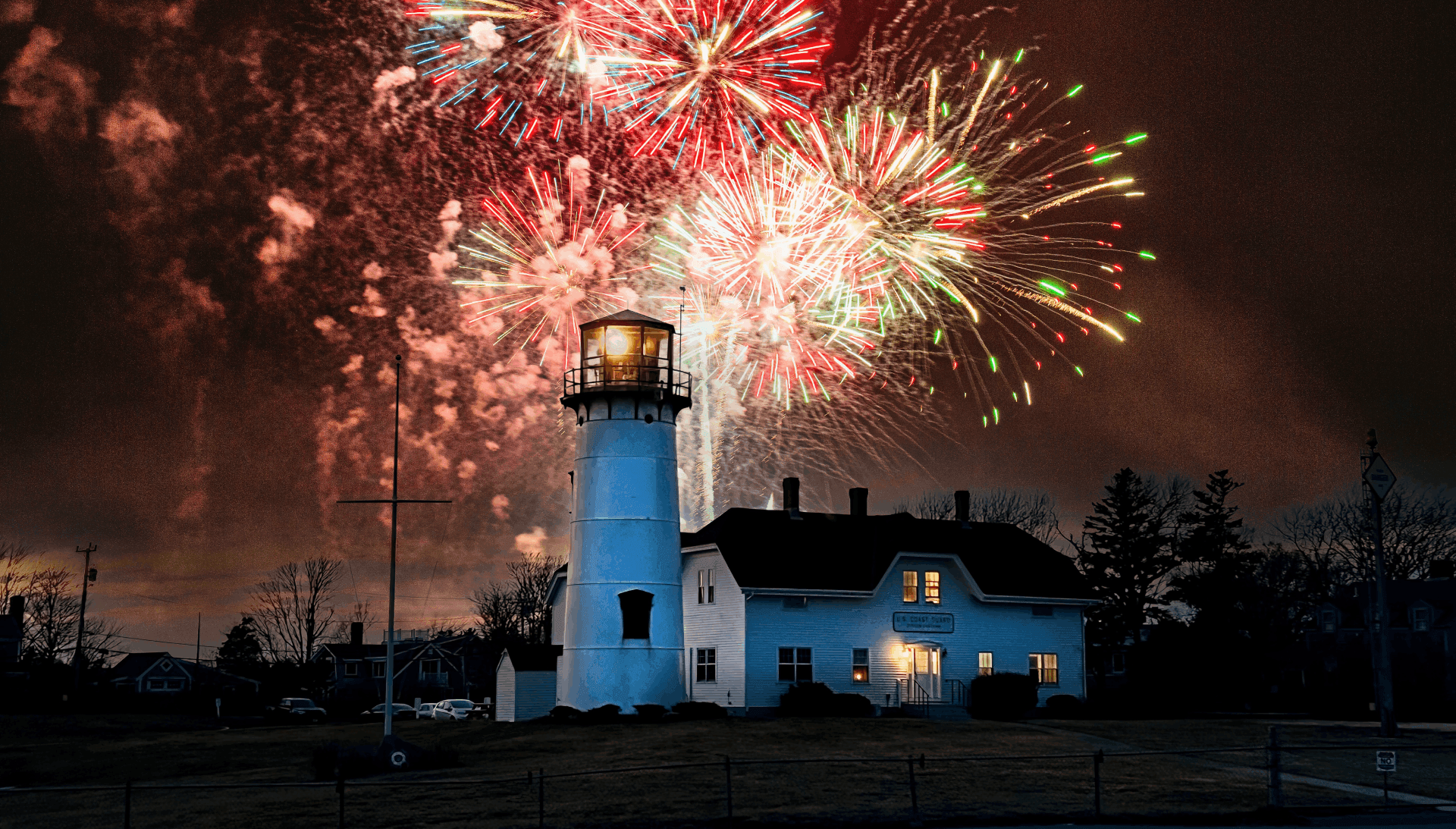 A lighthouse illuminates the night sky under a vibrant display of fireworks.