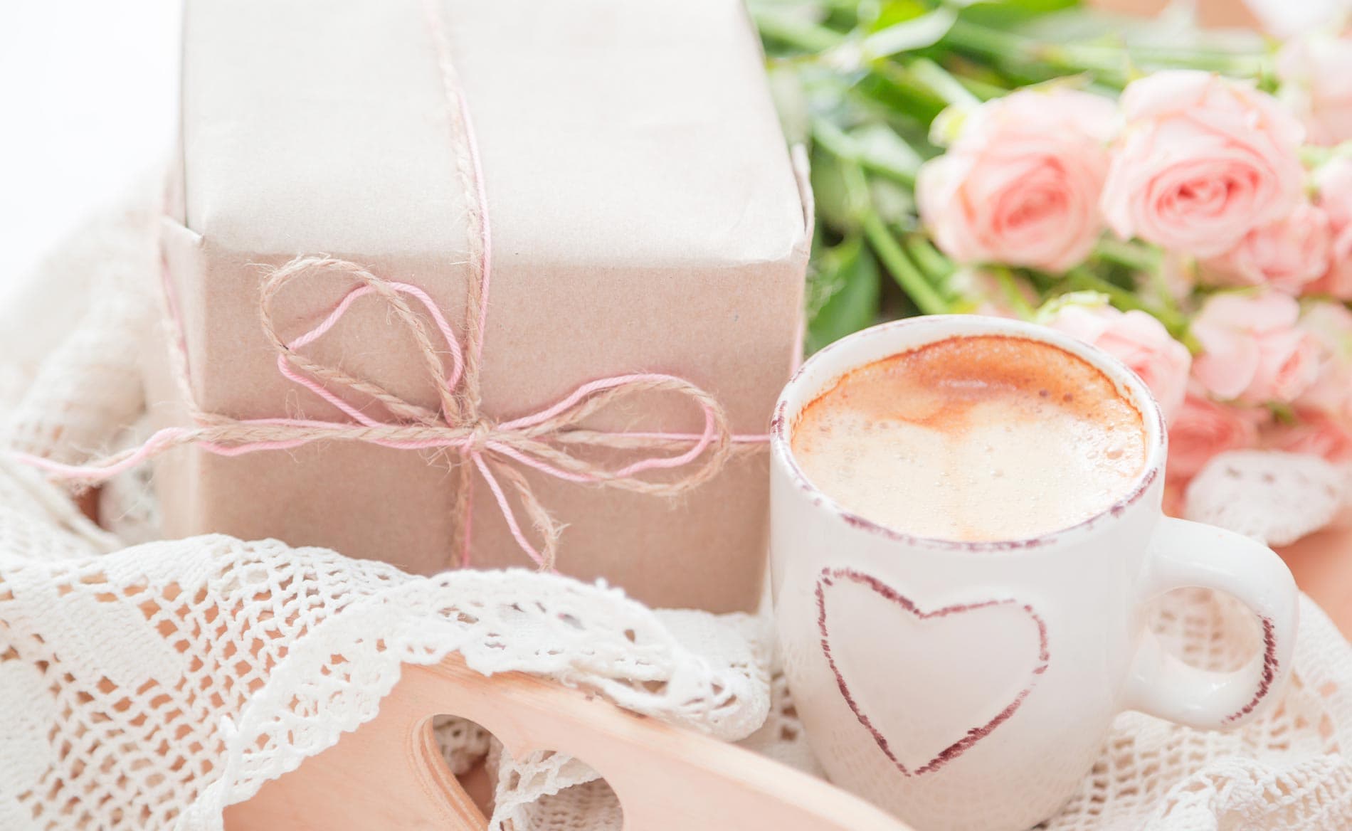 A cozy scene featuring a heart-decorated mug of coffee, a wrapped gift, and pink roses on a lace tablecloth.