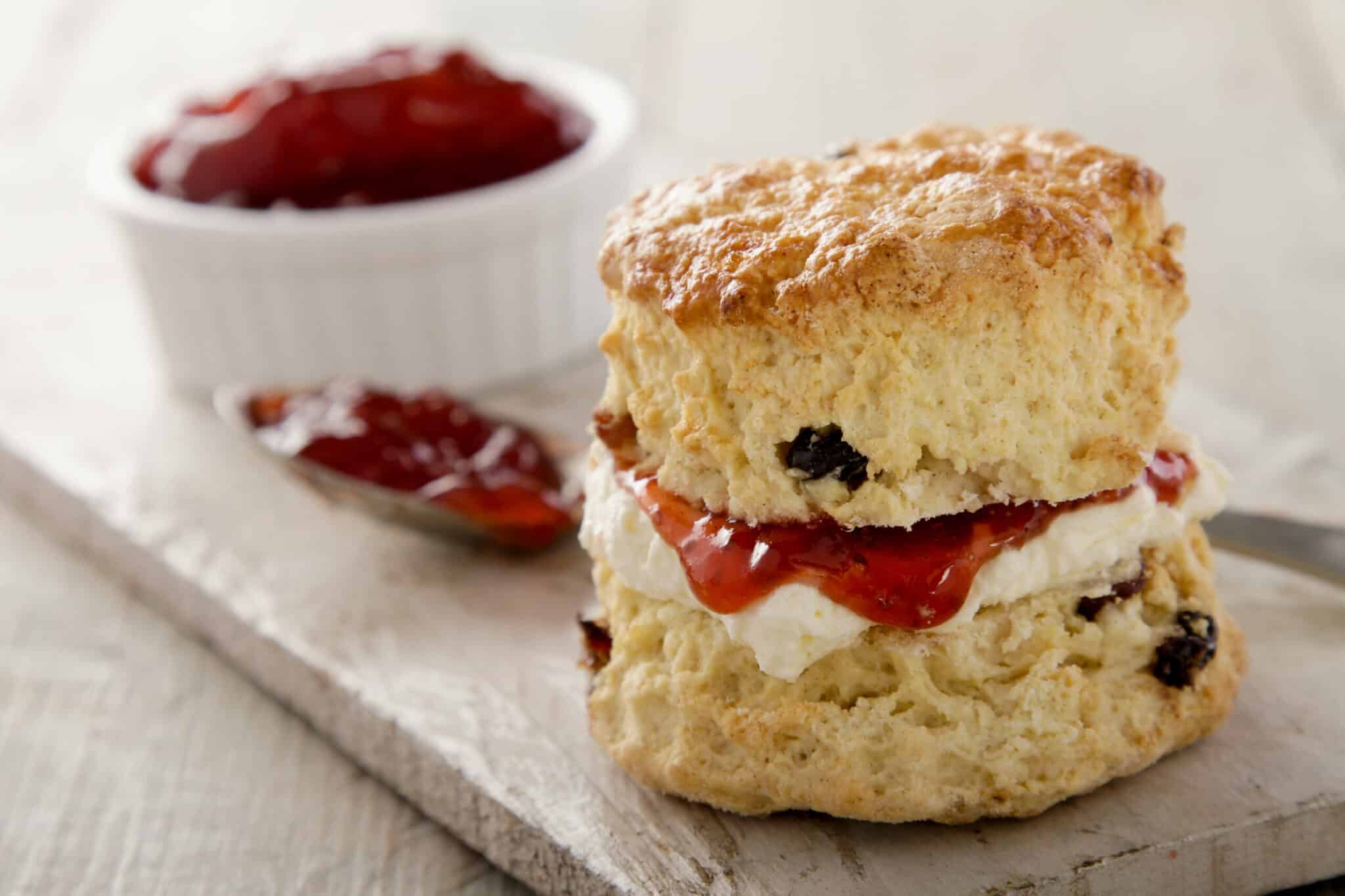 A freshly baked scone layered with cream and strawberry jam on a wooden surface.