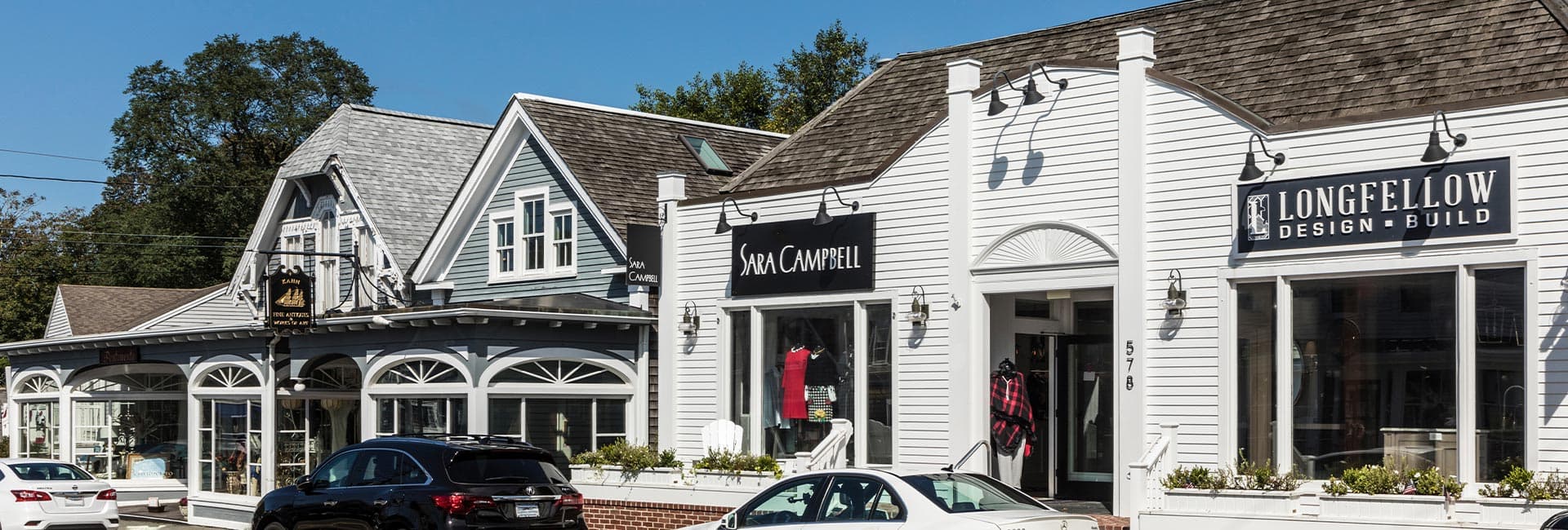 A row of quaint, white storefronts featuring a clothing shop and a design-build business, set against a clear blue sky.