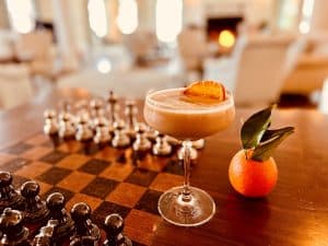 A cocktail garnished with an orange slice sits on a chessboard next to an orange.
