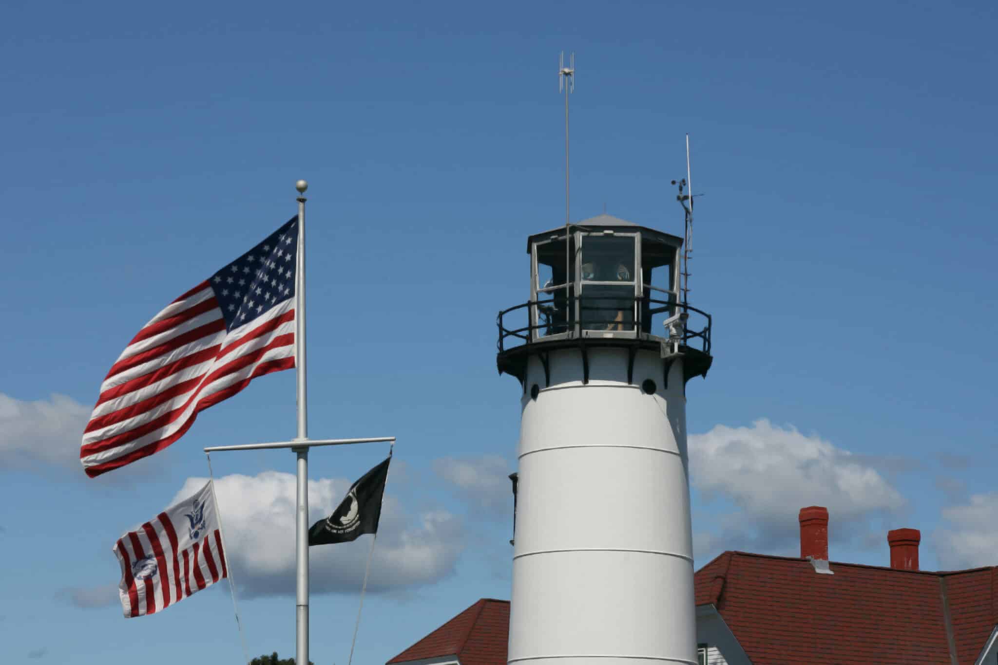 A lighthouse stands beside American flags against a clear blue sky.