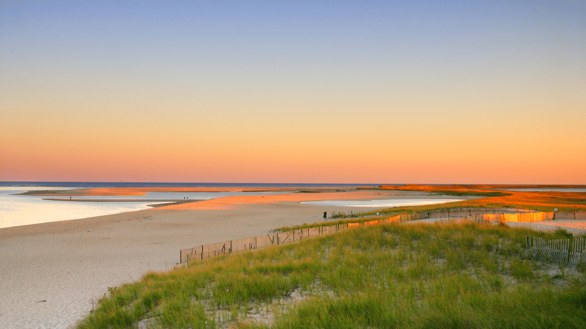 Sunset over a tranquil beach with sandy shores and grassy dunes.