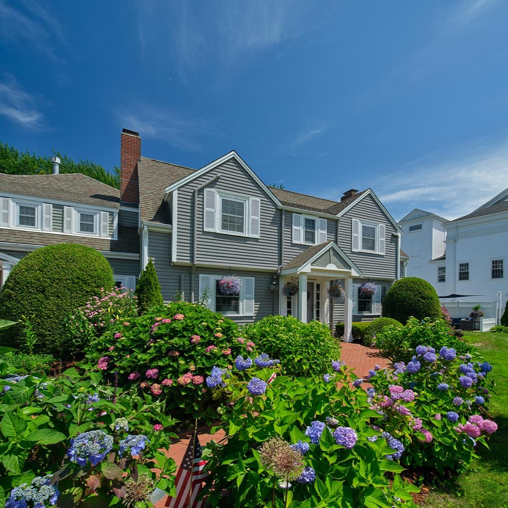 A charming gray house surrounded by lush green shrubs and colorful flowers under a clear blue sky.