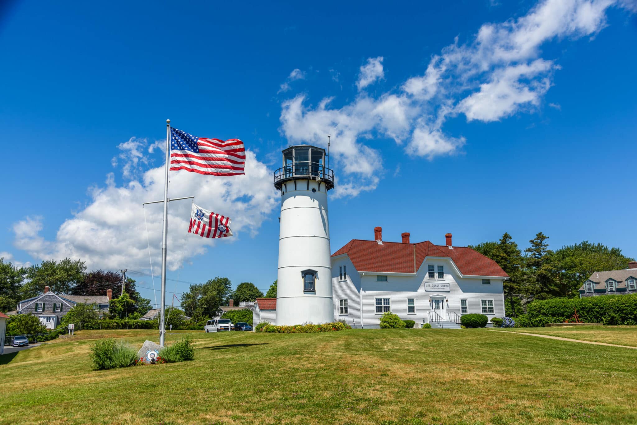 A white lighthouse stands beside two American flags against a blue sky with fluffy clouds.