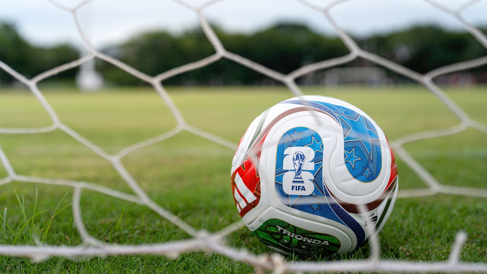 A close-up of a FIFA soccer ball resting on grass, framed by a goal net. A close-up of a FIFA soccer ball resting on grass, framed by a goal net.