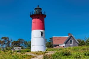 A red and white lighthouse next to a small house, surrounded by greenery under a clear blue sky.