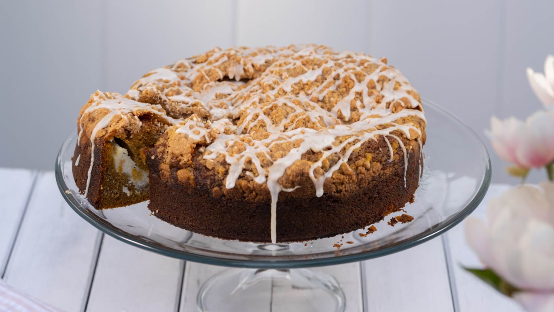 A moist coffee cake with a crumb topping and drizzle, displayed on a glass cake stand.