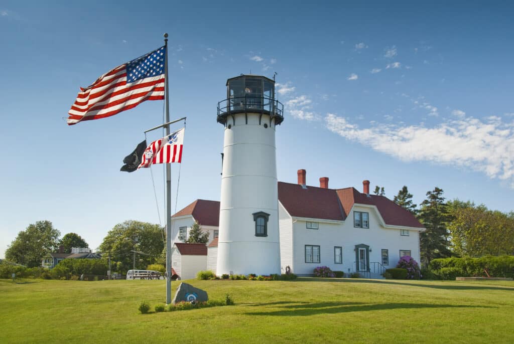 A white lighthouse with a red-roofed building beside it, featuring American flags in the foreground.