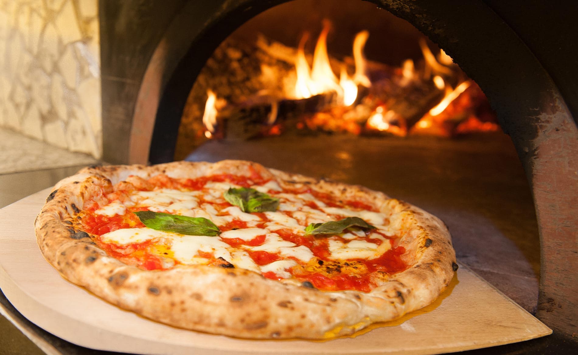 A freshly baked pizza with melted cheese and basil sits on a wooden board in front of a brick oven.