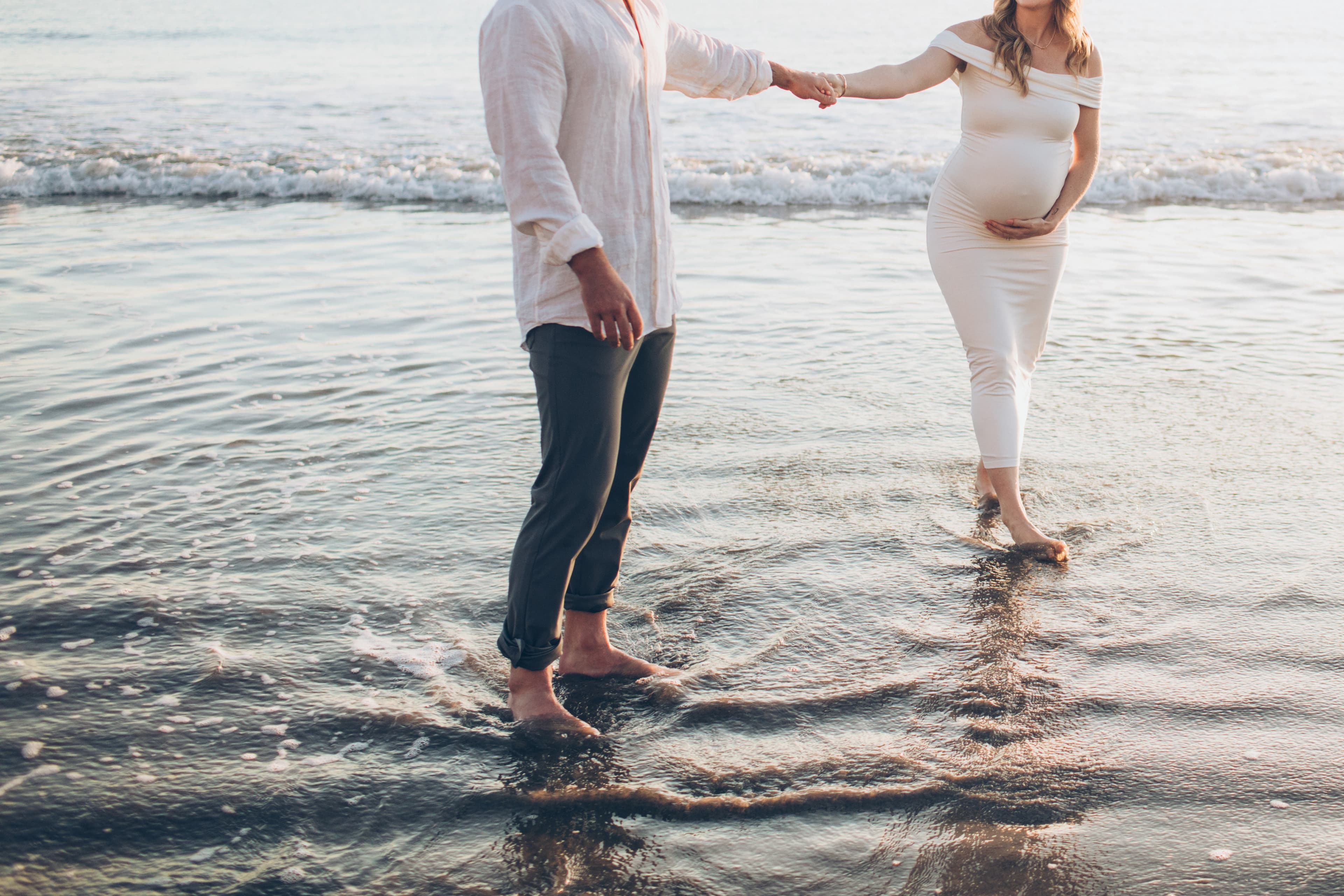 A pregnant woman and a man hold hands while standing in shallow water at the beach. A pregnant woman and a man hold hands while standing in shallow water at the beach.