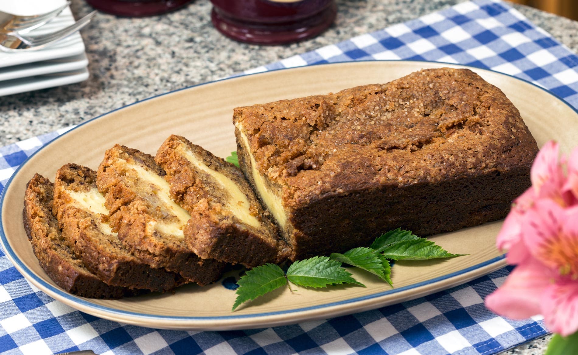 Sliced loaf of cake served on a platter with green leaves, set on a blue and white checkered tablecloth.