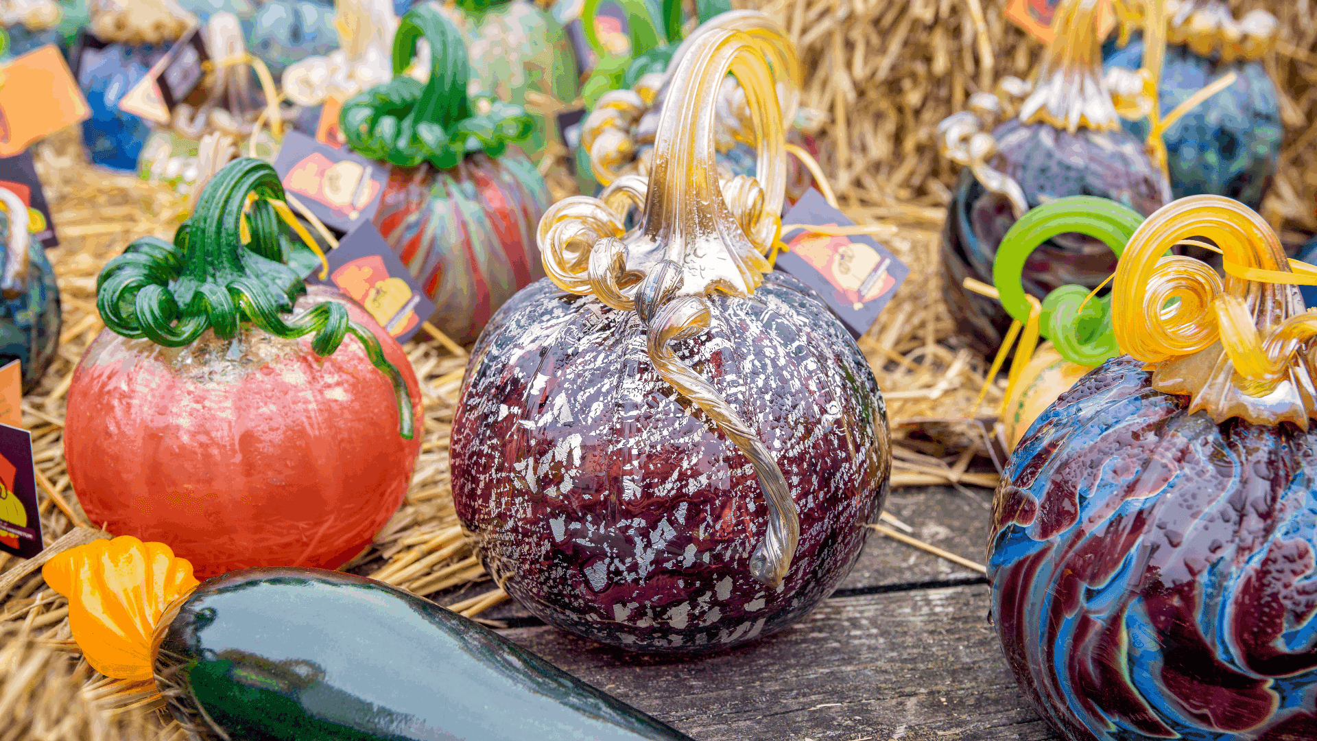 A collection of colorful glass pumpkins and vegetables displayed on a wooden surface surrounded by straw.